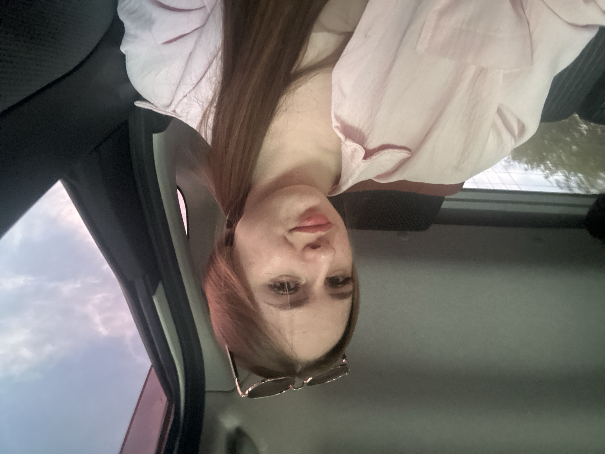 A close-up portrait features a young woman with long, straight brown hair, wearing a light pink collared shirt. Her face is in the foreground, with sunglasses perched on her forehead. She looks directly towards the camera with a neutral to slightly pensive expression, her lips slightly parted.
The setting appears to be inside a car, with elements of the vehicle's interior visible around her, including a dark headliner and a side pillar. Through the windows, a cloudy or overcast sky is visible, along with hints of green foliage from trees, suggesting a daytime scene. The overall lighting is soft and diffused. No text is visible in the image.