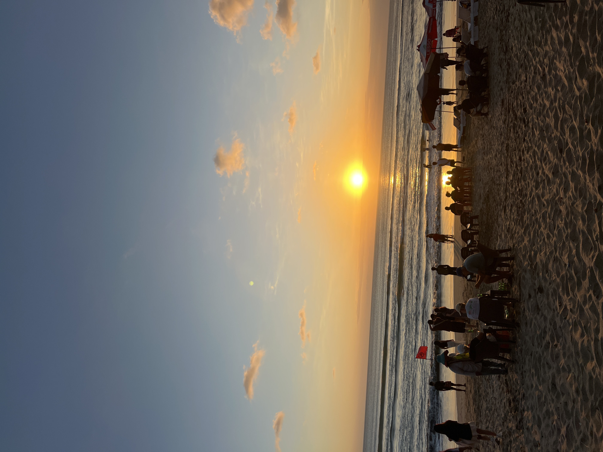 A beach scene in Kuta, Indonesia, at sunset. The sun is low on the horizon, casting a warm orange glow across the sky and water. Gentle waves roll onto the sandy shore. Silhouettes of people are scattered across the beach, some standing, some sitting in chairs, and some appearing to be walking. Several beach umbrellas are visible, along with lounge chairs. A red flag is planted in the sand near the water's edge. The overall atmosphere is relaxed and peaceful.