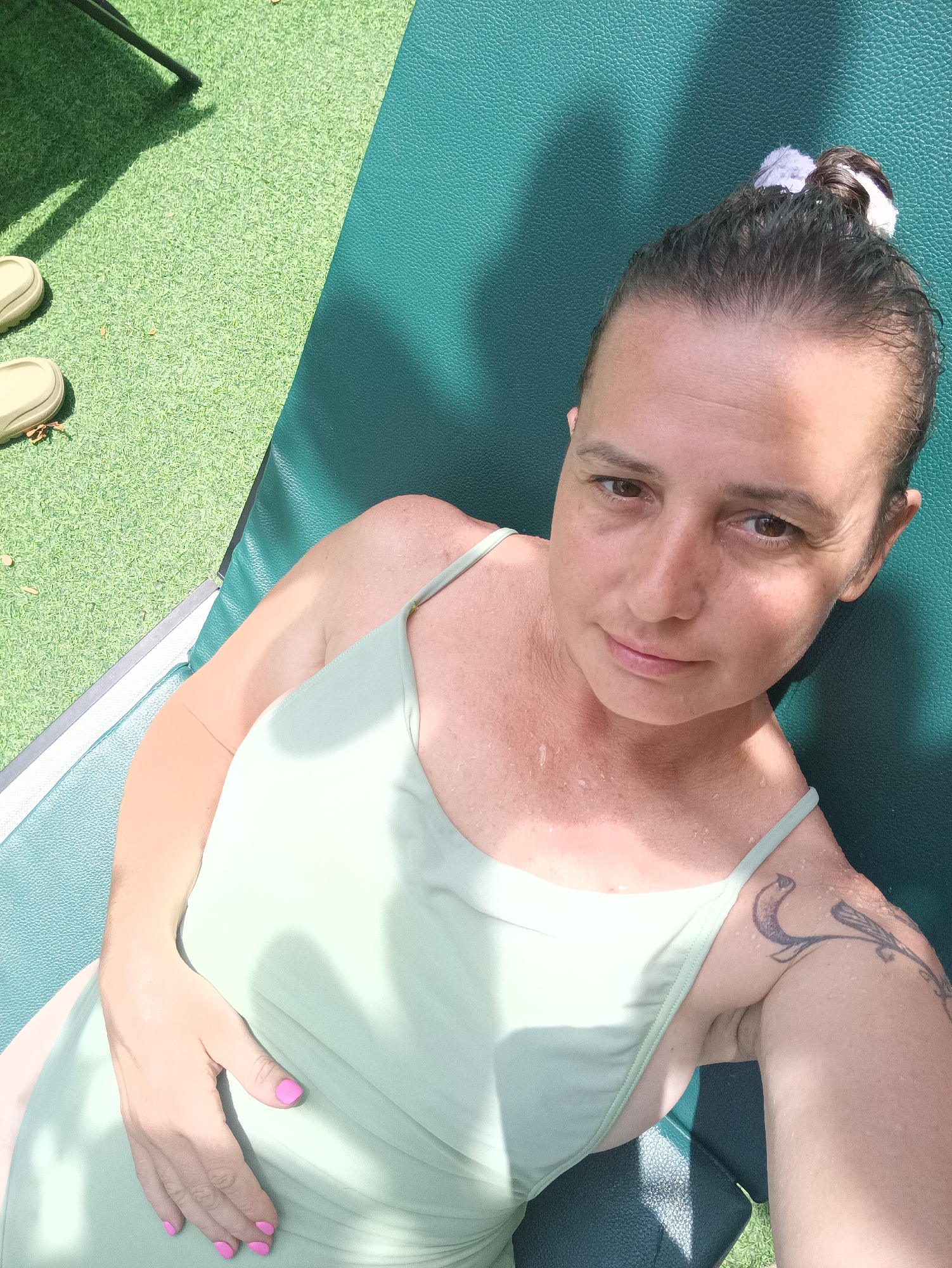 A woman is lying on a green chaise lounge outdoors on a sunny day. She is wearing a light green swimsuit and has her hair pulled back with a white hair tie. She has a tattoo on her left shoulder. The lounge is situated on a green artificial turf surface. A pair of sandals are visible on the turf. The location is Hua Hin City Municipality, Thailand.