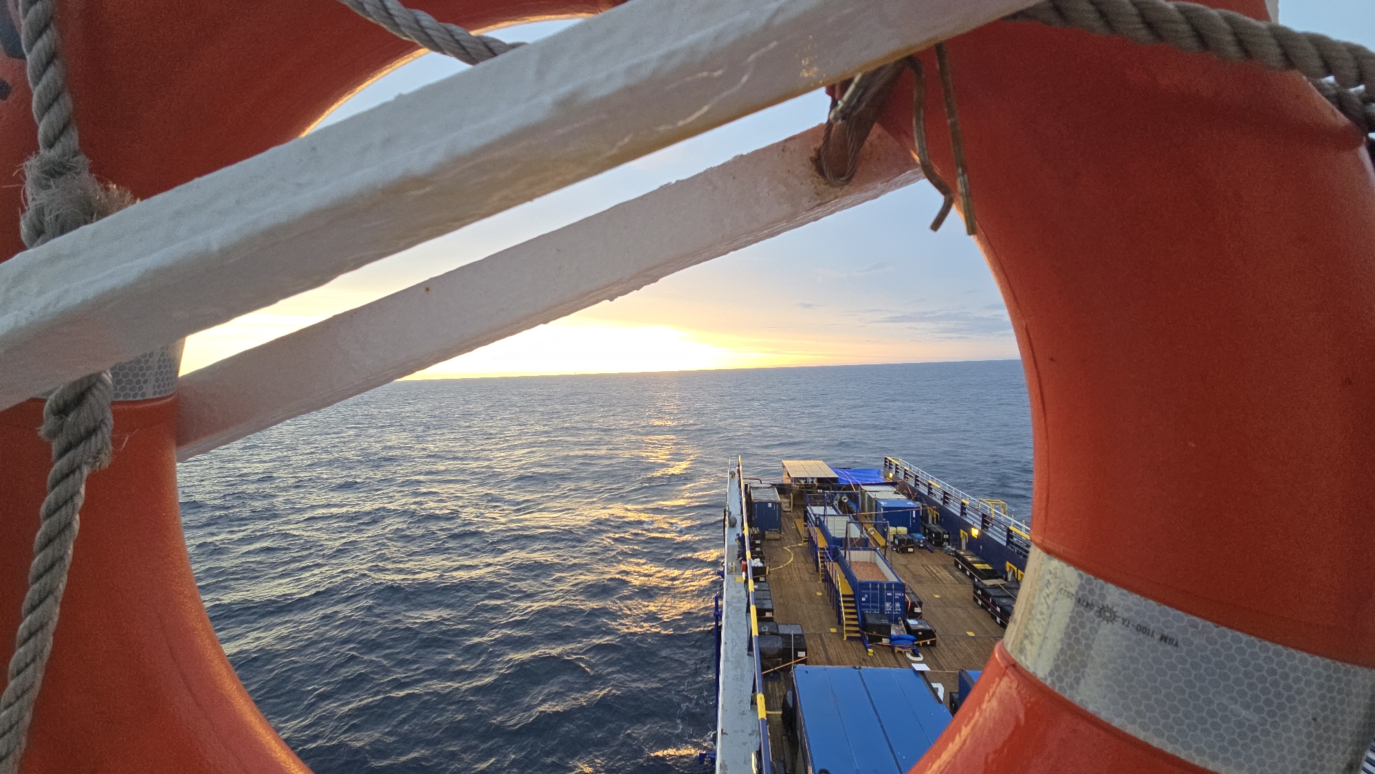 A view from the deck of a ship, looking towards the ocean at sunset. The foreground includes parts of an orange life preserver with a reflective strip and a white railing, framing the view. The ocean occupies the majority of the image, with sunlight reflecting off the water's surface. The deck of the ship is visible towards the bottom of the frame, revealing containers and equipment. The location is likely at sea, with no land or city visible.