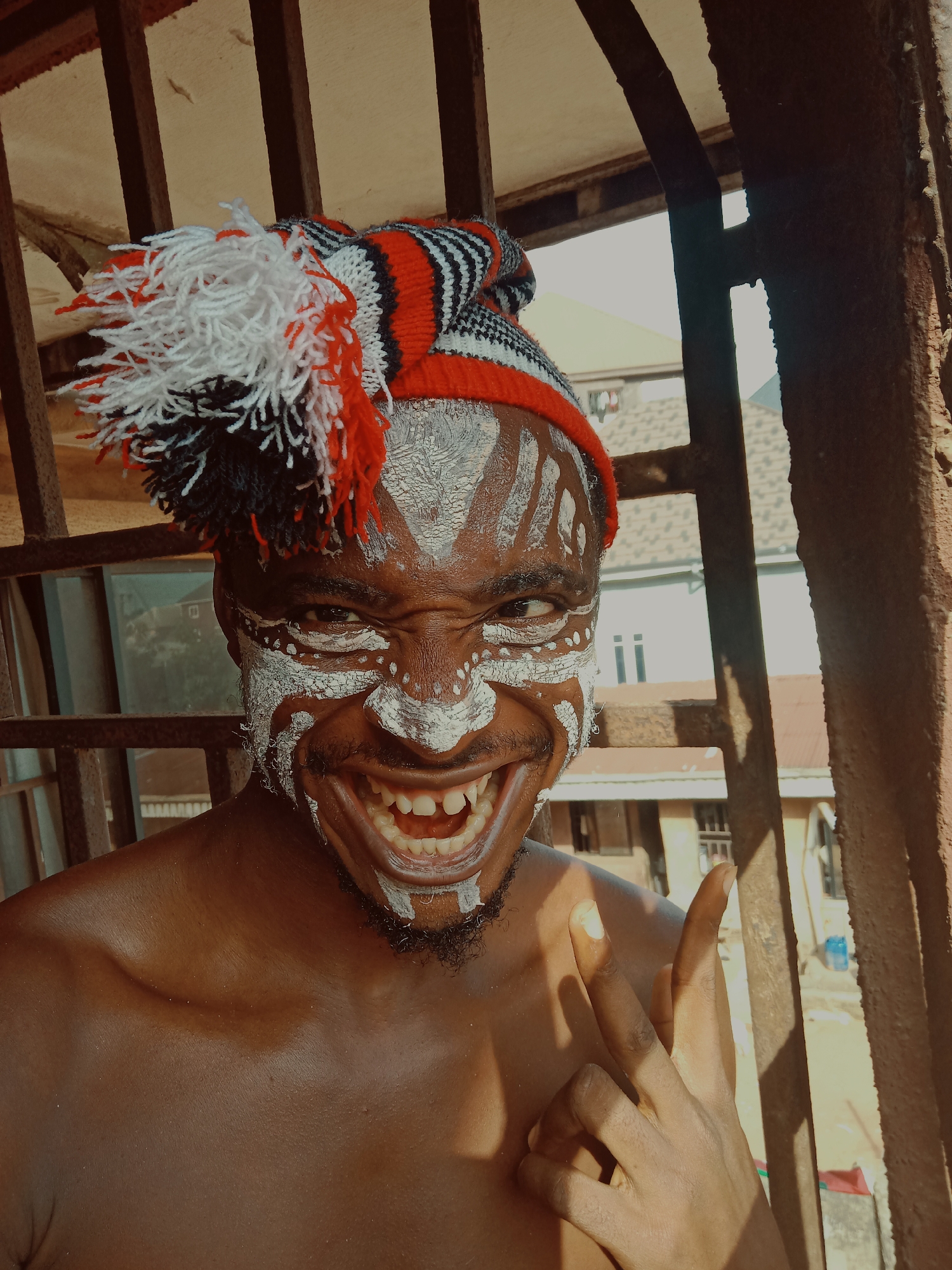 A medium, eye-level shot depicts a man, identified as being in Nkpor, Nigeria, with his upper torso exposed. The man's face is adorned with white paint in a tribal pattern, and he wears a red, white, and black striped knit cap with a large pompom on top. He is grinning broadly, exposing his teeth, and his facial expression is intense. His right hand is raised, forming a "devil horns" hand gesture.
The scene appears to be outdoors, as he is standing near a rusted metal railing and a window. Behind him, glimpses of buildings and structures are visible, confirming the urban location. The lighting suggests daylight.