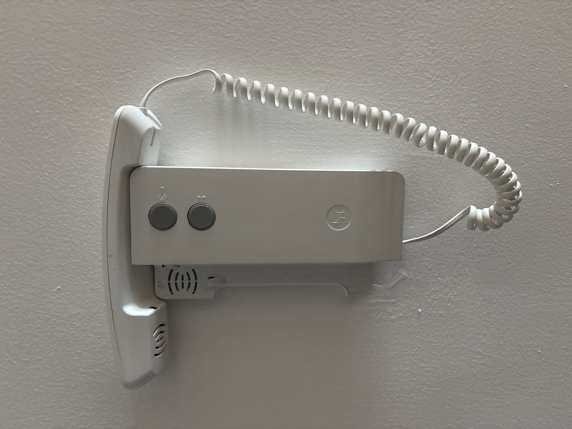 An off-white intercom unit is mounted on a textured, off-white wall. The intercom has a handset that rests on a base. The handset is slightly elevated, suggesting it is not currently in use. Two circular buttons are visible on the front of the unit. A coiled, off-white cord connects the handset to the base. A circular logo with the letters "bpt" is present on the lower section of the intercom unit. The wall exhibits some minor imperfections, such as small specks of dirt or debris. The lighting suggests an indoor environment. This object is likely a common fixture in residential or commercial buildings in Dubai, United Arab Emirates, used for communication within the building or with external visitors.