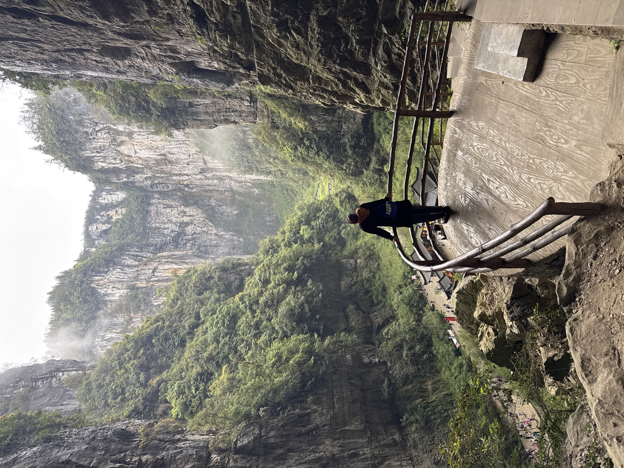 A person stands on a wooden walkway with a wooden railing, overlooking a vast, green landscape, framed by towering rock formations. The location is the Wulong District in China. The rock formations are partially covered in lush vegetation and disappear into the distance. The bottom of the valley shows traces of human activity including buildings and people. The viewpoint is at a high elevation. The overall scene conveys a sense of grandeur and natural beauty.