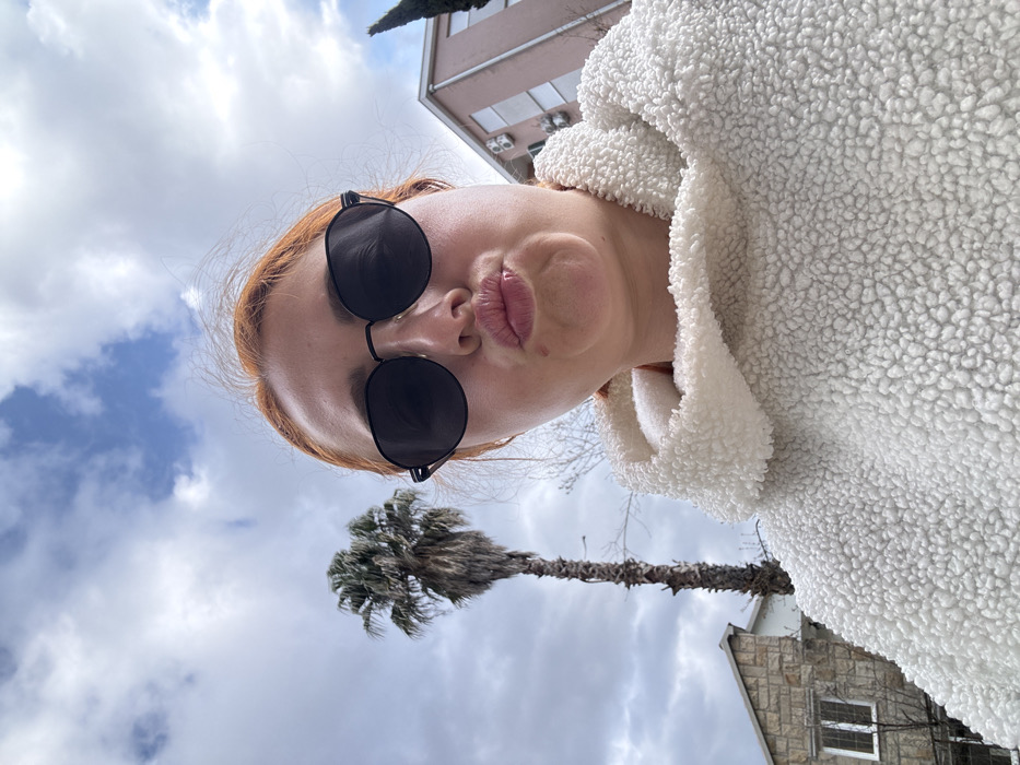 An upward-angle selfie captures an adult female outdoors. The subject, in the foreground, has red hair and wears dark, octagonal sunglasses, pouting her lips. She is dressed in a white, textured, hooded garment.
The background reveals a partially cloudy sky. To the subject's left, a tall, slender palm tree with a visible trunk and sparse fronds stands. Below the palm, a building with stone cladding and a white-framed window is partially visible. To the subject's right, a multi-story building with a light pink facade and multiple external air conditioning units is present. A dark, conical evergreen tree is visible behind the pink building.
The environment depicts an urban-coastal residential area consistent with the architecture and vegetation found in Budva, Montenegro.