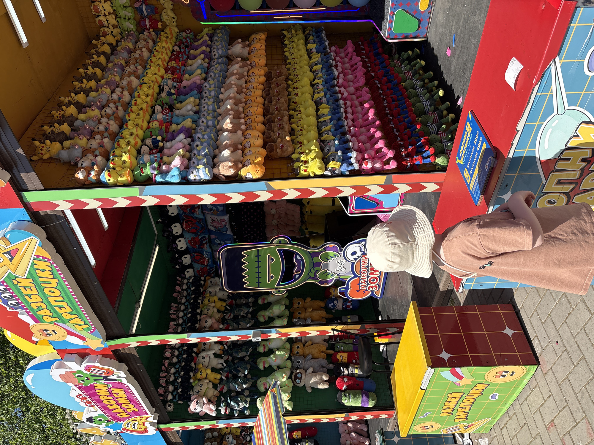A child stands near a "shoot the target" game stall in Izhevsk, Russia. The stall has multiple levels of stuffed animals as prizes. The stall has signs in Russian and is decorated with cartoon characters. The child is wearing a white hat and brown shirt.