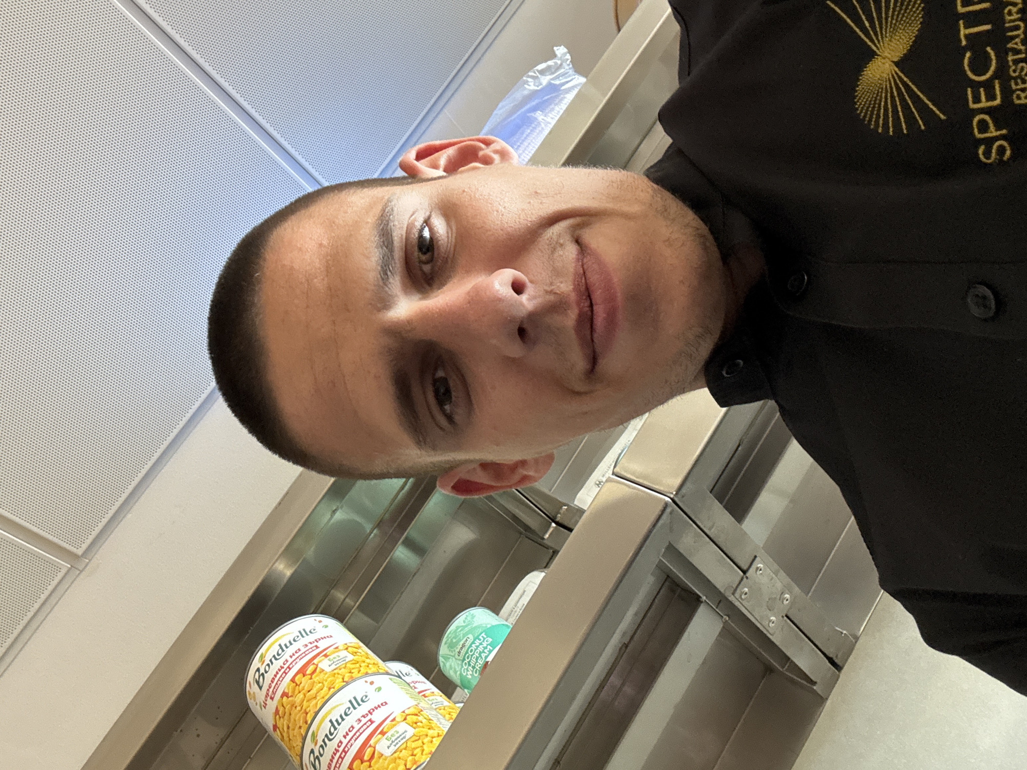 A man wearing a black chef's uniform with the "Spectra Restaurant" logo is taking a selfie in what appears to be a kitchen. The background shows shelves holding canned goods, including "Bonduelle" corn and "dragon" coconut whipping cream. The location is potentially in Sofia, Bulgaria, based on the Cyrillic text on the "Bonduelle" cans. The lighting is bright, suggesting an institutional or commercial setting. The man is tilting his head slightly and looking into the camera with a slight smile. The perspective is from above, capturing the man and some of the shelving.