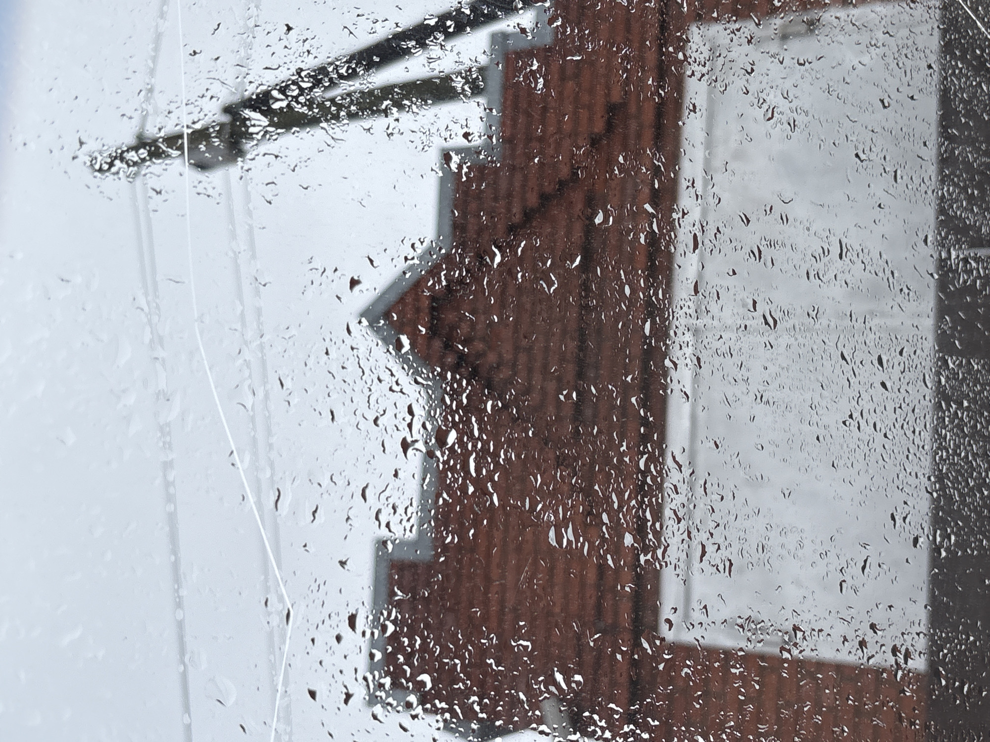 A rainy day view of a building through a wet window. The building appears to have a stepped facade and is brick. A metal fixture, possibly an antenna, is visible at the top left. It is not possible to confirm the location is in the city of Прикамский, Russia.