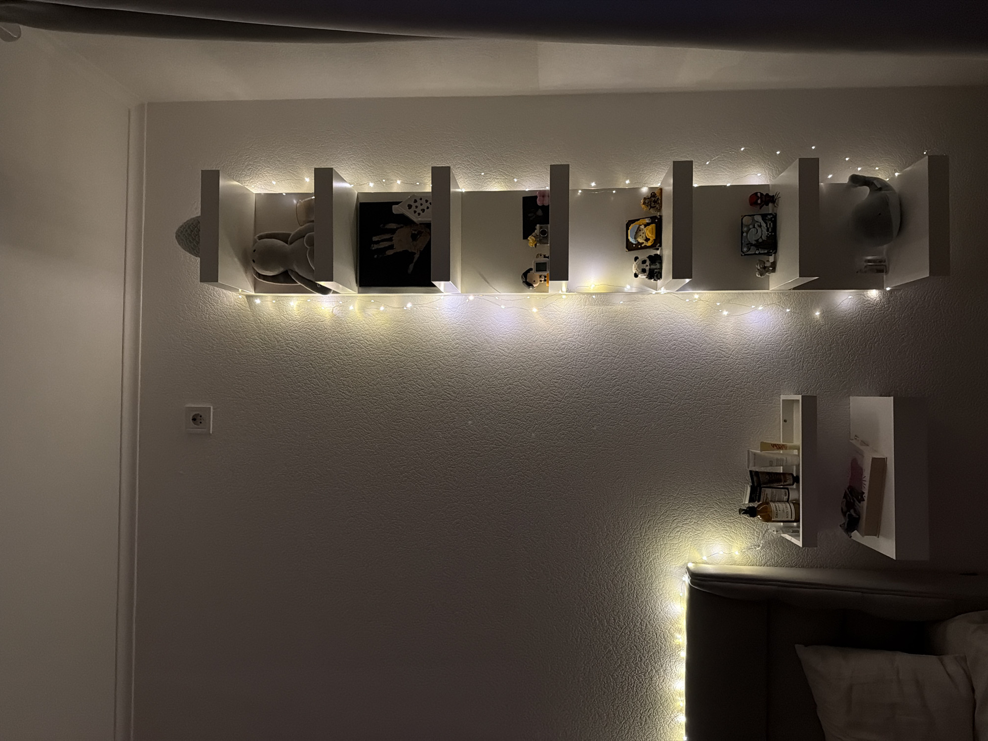 The image depicts a bedroom scene, likely in Kyiv, Ukraine. The focal point is a white, vertical shelving unit mounted on a textured white wall. The shelves are decorated with a string of warm-toned LED fairy lights that trace the perimeter of the unit.
On the shelves, various decorative items are displayed. The top shelf holds a small grey decorative object, possibly a knitted egg or shell, and a large, soft stuffed rabbit. The shelf below it contains a framed print of a handprint, a small white house-shaped object, and a white vase with small decorative elements. Further down, one shelf displays small figurines, including what appears to be a panda, a person in yellow, and a brown bear. Another shelf holds a small elephant figurine, a square object with a graphic design, and a red figurine of what resembles Spider-Man. The lowest shelf has a grey whale-shaped plush toy.
To the left of the vertical shelf, near a headboard of a bed, there are two horizontal, floating shelves. The upper shelf holds several bottles and tubes of cosmetic products. The lower shelf displays a book. The wall is illuminated by the fairy lights and appears to have an electrical outlet near the top. A grey curtain is visible on the far right of the image. The lighting in the room is dim, with the fairy lights providing the primary illumination.