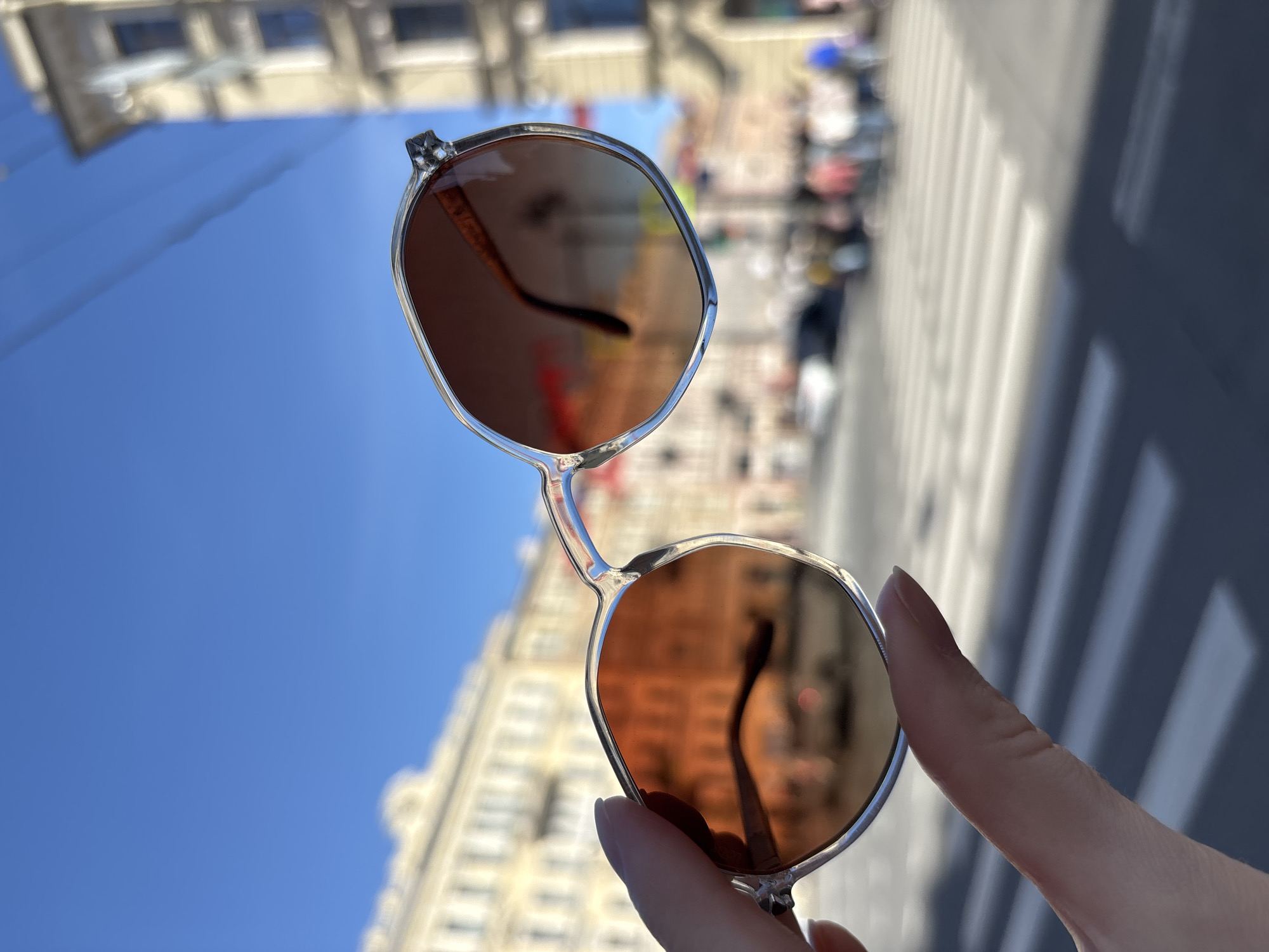The image depicts a hand holding a pair of brown-tinted sunglasses against a blurred backdrop of Saint Petersburg, Russia. The foreground focuses on the silver-framed sunglasses, showcasing reflections of the surroundings in the lenses. The background reveals a street scene with blurred pedestrians, buildings, and a pedestrian crossing. The sky is a bright blue, visible in the upper left corner of the frame. The hand holding the sunglasses is fair-skinned. The scene appears to be captured during daylight hours, with a clear, sunny sky contributing to the overall bright lighting.