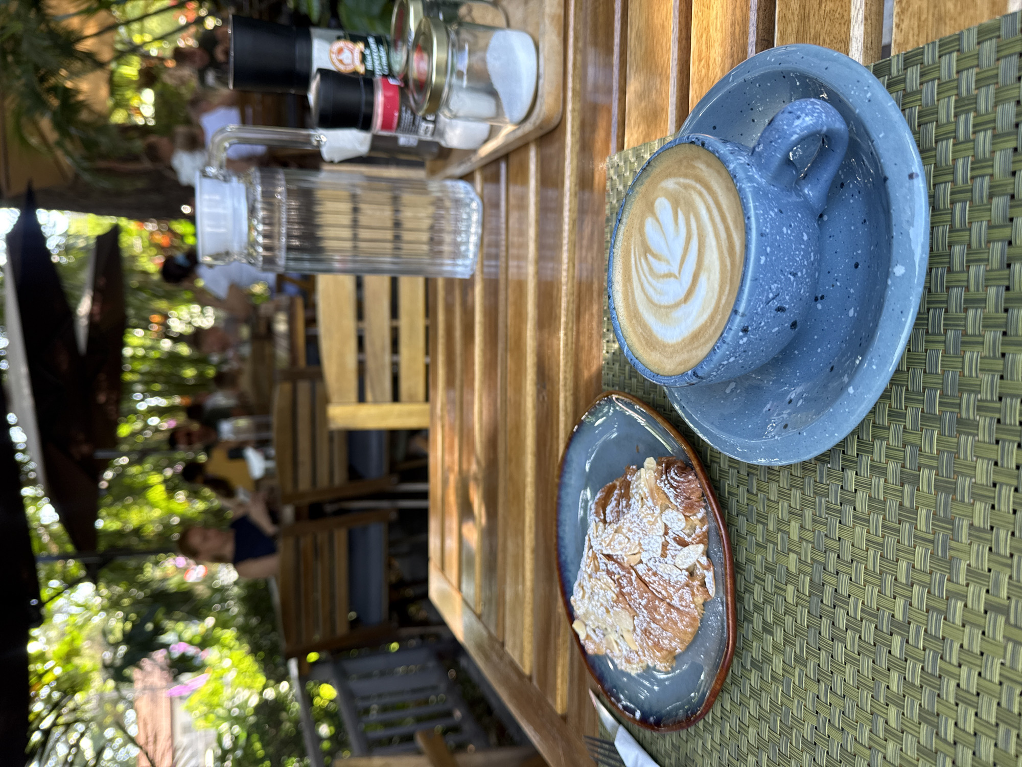 The image captures a close-up view of a wooden table at an outdoor cafe in the Ko Samui District of Thailand. In the foreground, a blue speckled ceramic cup filled with latte art sits on a matching saucer. Beside it, on a gray plate, is a flaky croissant dusted with powdered sugar and sliced almonds. To the right, a clear glass pitcher of water, a salt and pepper shaker set, and a small jar of sugar are arranged on a wooden tray. The background is blurred, but shows people seated at tables under umbrellas, with lush greenery and palm trees suggesting a tropical setting. The overall impression is a relaxed dining experience in a pleasant outdoor environment.
