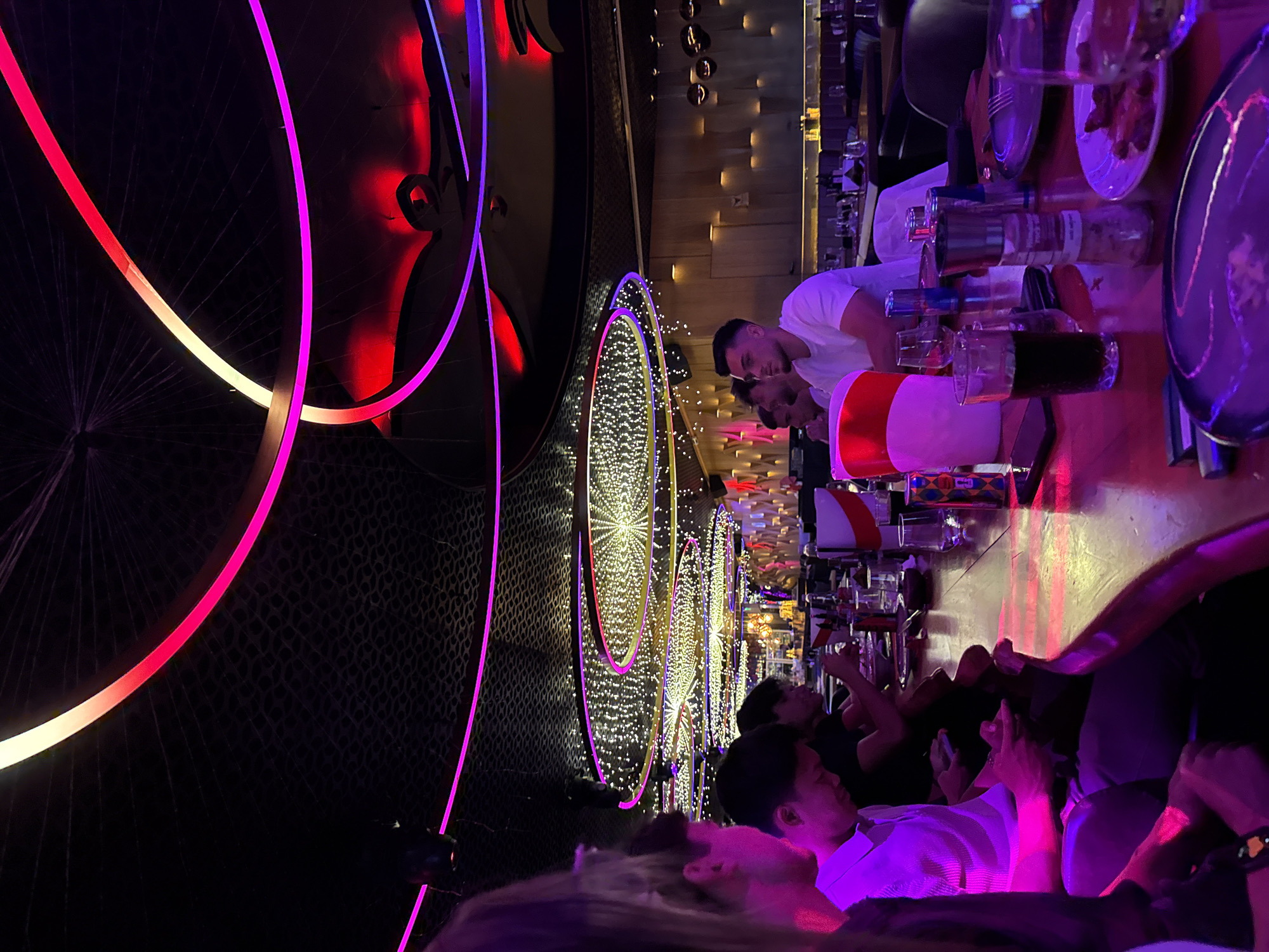 The image depicts the interior of a dimly lit, modern restaurant or bar, likely located in Sofia, Bulgaria, characterized by its vibrant neon lighting and contemporary decor. In the foreground, four individuals are seated at a bar, illuminated by the ambient pink and purple neon lights. The person on the far left is partially visible, with their back to the camera, their dark hair and shoulder suggesting they are female. To their right, a man in a white polo shirt is looking down at his phone, his face in shadow. Next to him, another man, also with dark hair, is looking down, possibly at his phone as well. The fourth person, a man with short dark hair, is wearing a white t-shirt and looking forward.
The bar itself is polished and reflective, showing the neon glow and the items placed upon it. These include glasses, bottles, a salt and pepper grinder, and plates with food. In the background, other patrons are visible at tables, and the restaurant's ceiling features an intricate design with multiple circular light fixtures adorned with small white lights, creating a star-like effect. The walls are adorned with textured panels and strategically placed lighting, contributing to the sophisticated and energetic atmosphere. The overall scene suggests a lively evening out, with patrons enjoying drinks and conversation in a stylish urban setting.
