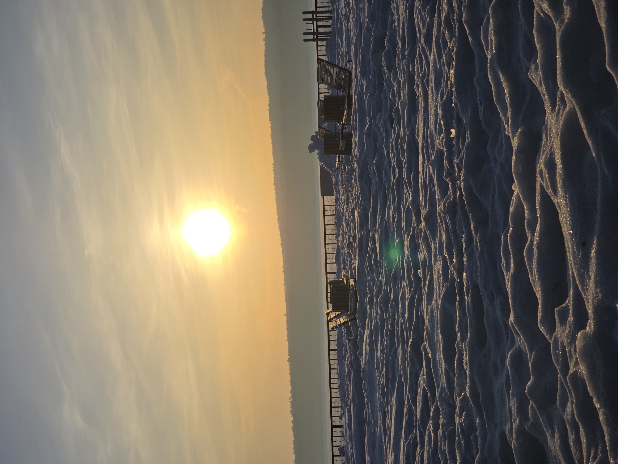 The image captures a winter scene at sunset in what could be Chaikovsky, Russia. The sun, a bright disc in the sky, casts a warm, golden light across the landscape. The foreground features a snow-covered area with pronounced ridges and patterns. A metal fence runs parallel to the beach, and a few wooden beach chairs are visible behind it. In the distance, a tree line or hill is visible, silhouetted against the bright sky. The sky shows a gradient of colors, transitioning from light blue to a warm yellow near the sun. The orientation of the image is vertical.