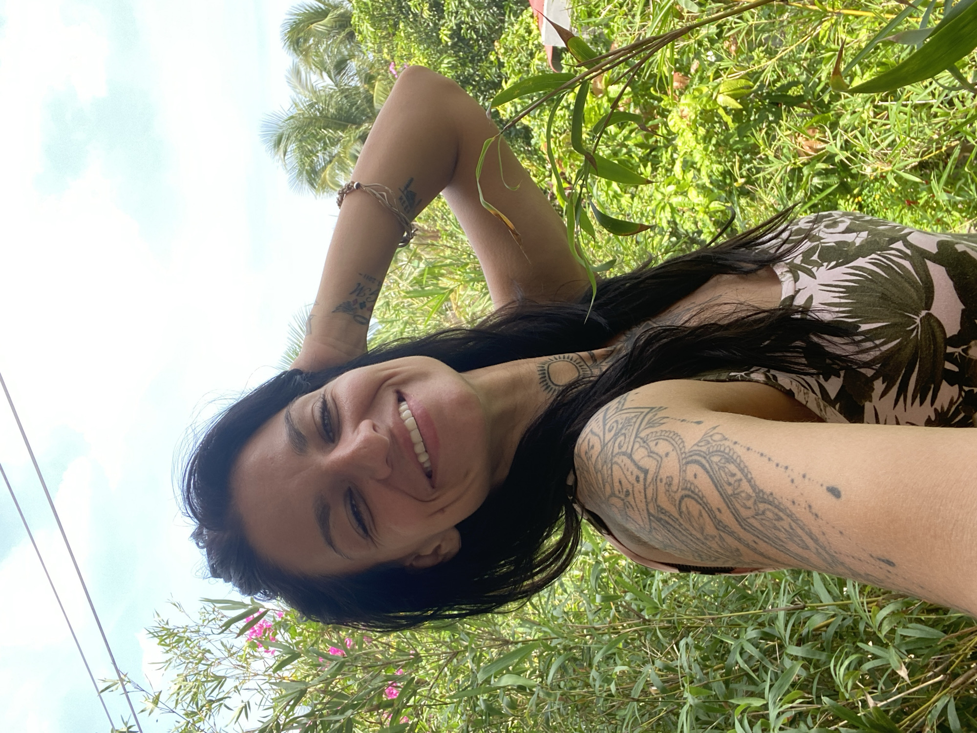 A woman with dark hair and tattoos on her arms and neck smiles at the camera, holding her arm behind her head. She is wearing a light-colored top with a tropical pattern. The background is lush green foliage, with some pink flowers visible. In the distance, palm trees can be seen against a blue sky with white clouds. This scene is set in Mũi Né, Vietnam, where the abundant vegetation and palm trees are characteristic of the region.