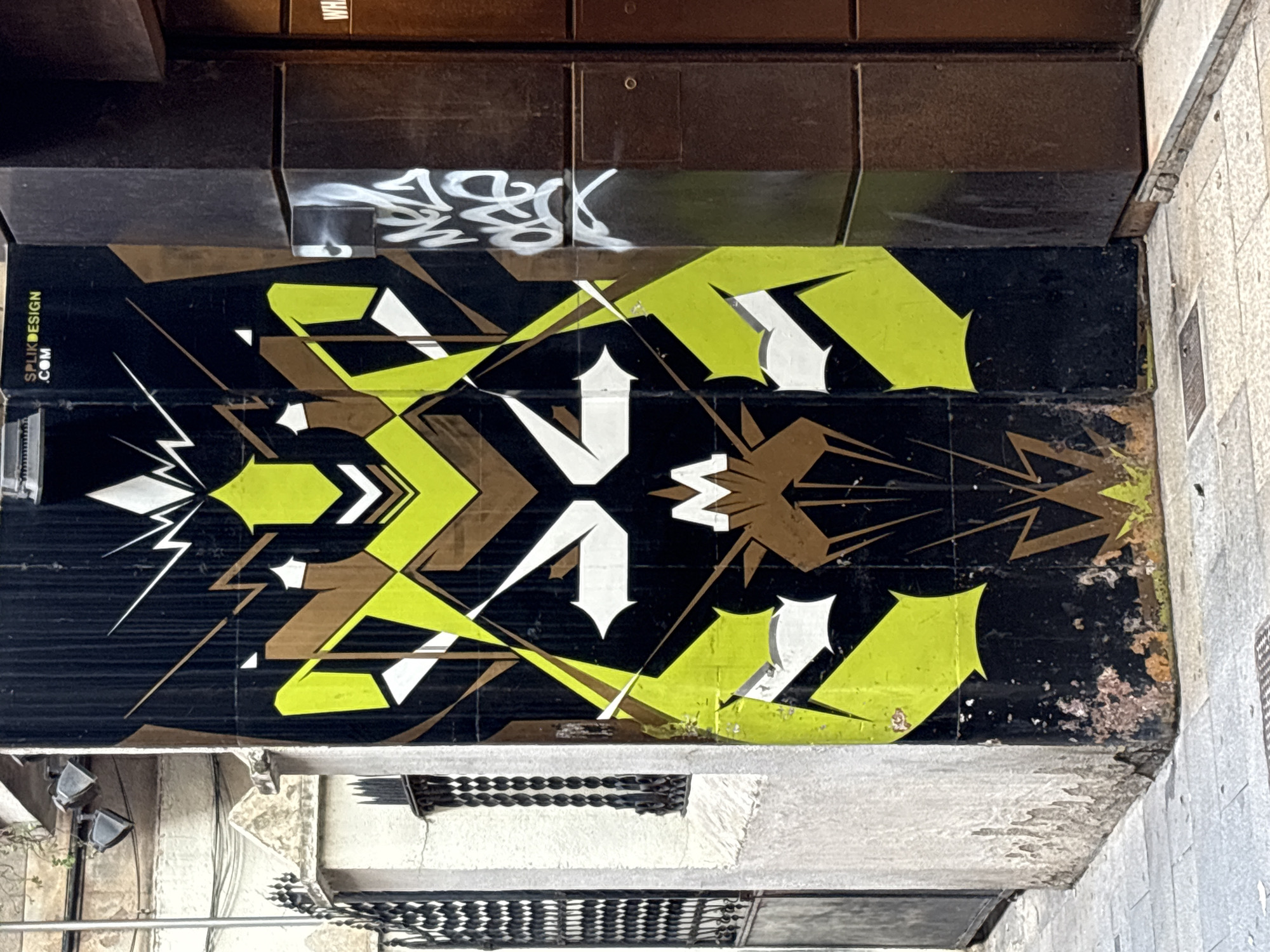 The image shows a section of wall in Barcelona, Spain, covered with black, green, white, and brown graffiti. The design features geometric shapes, arrows, and lightning bolts. The upper portion of the wall is made up of dark brown panels with some white graffiti. A website address "SPLIKDESIGN.COM" is visible on the left. Below the graffitied section, there's a lower wall with metal grates or air vents. The setting appears to be an alleyway or narrow street. The environment consists of stone walls, metal grates, and a possibly aging building façade.