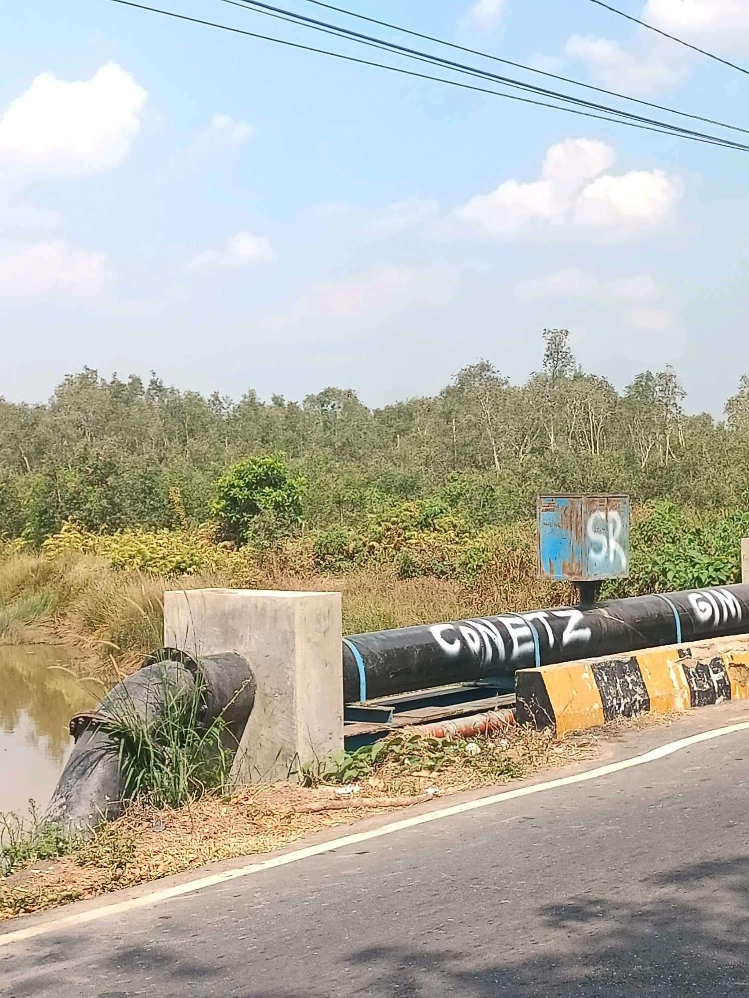 In the city of Sungai Rutas Hulu, Indonesia, a large black pipe is visible, exiting a concrete structure and extending horizontally across the frame. The pipe is supported by a concrete pillar and is part of an apparent infrastructure project. The pipe itself has white lettering, reading "CONETZ GIN" with a blue band. To the right of the pipe, a blue metal box with the letters "SR" is mounted on a post. The scene is situated outdoors, with a body of murky water to the left and a dense green forest bordering the background. The ground is covered in dry vegetation and soil, with a paved road in the foreground. Overhead, power lines are strung across the sky, which is a clear blue with scattered white clouds. The overall impression is a functional, perhaps industrial, setting within a natural landscape.