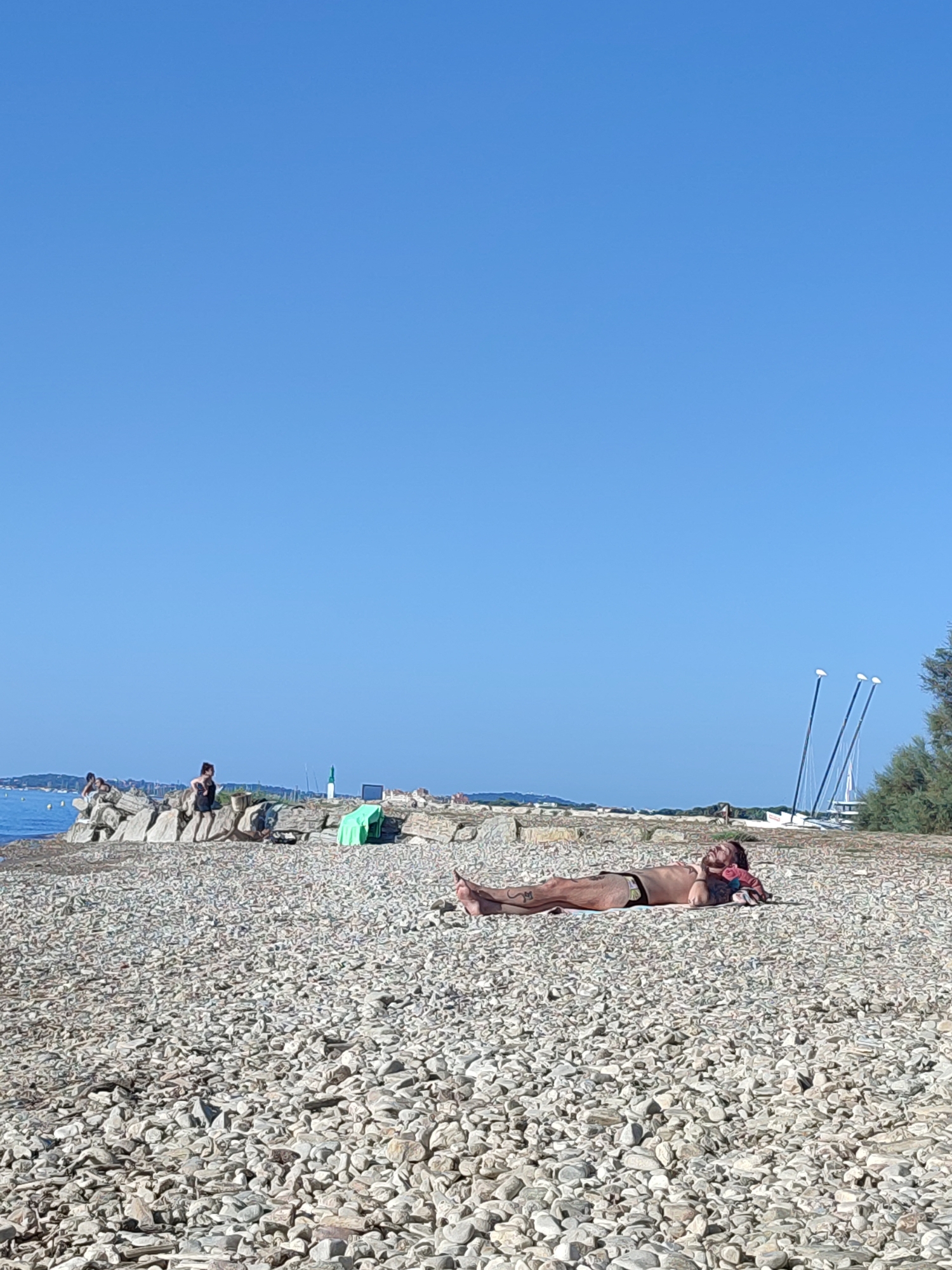 This image depicts a sunny day at the beach in Hyères, France. The foreground is dominated by a vast expanse of grey and white pebbles. In the middle ground, a man with tattoos is lying on his back on the pebble beach, facing upwards towards the clear blue sky, seemingly sunbathing. He is wearing dark swim trunks and has a towel or piece of clothing tucked under his head. To the left, a rocky embankment is visible, with several individuals sitting and standing on it. A body of water can be seen beyond the embankment. In the background, there are some small boats and trees, with the faint outline of buildings on the horizon. The overall atmosphere is one of leisure and relaxation on a warm summer day.