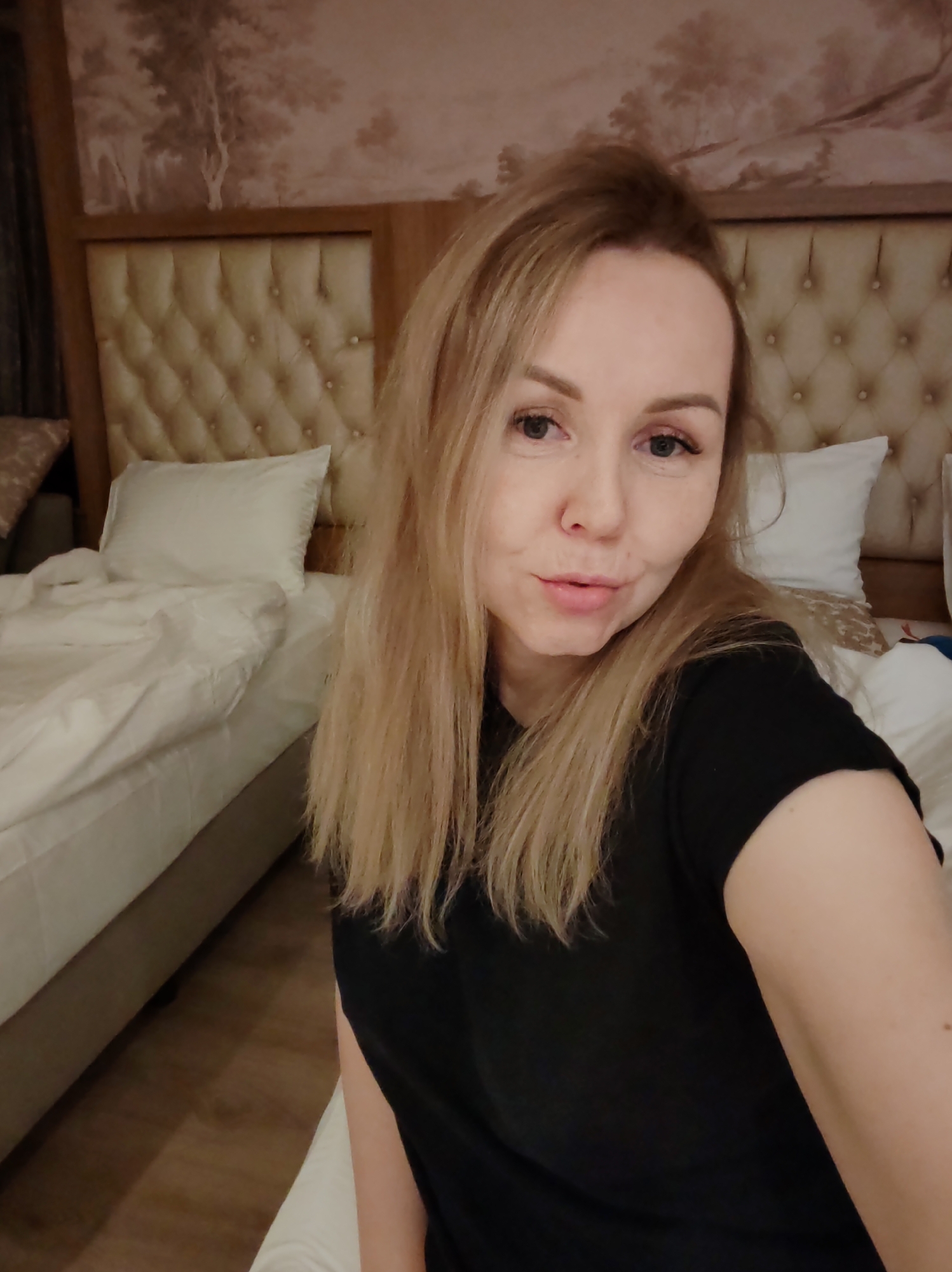 A woman with blonde hair, wearing a black t-shirt, is shown in a selfie. She has a fair complexion, and is posing with slightly pursed lips. She appears to be indoors, sitting on or near a bed. Behind her are two beds with white linens and quilted headboards. Above the headboards is a decorative panel featuring an image of trees and buildings in a brown-toned color scheme. The location is likely Alanya, Turkey given the type of interior design.