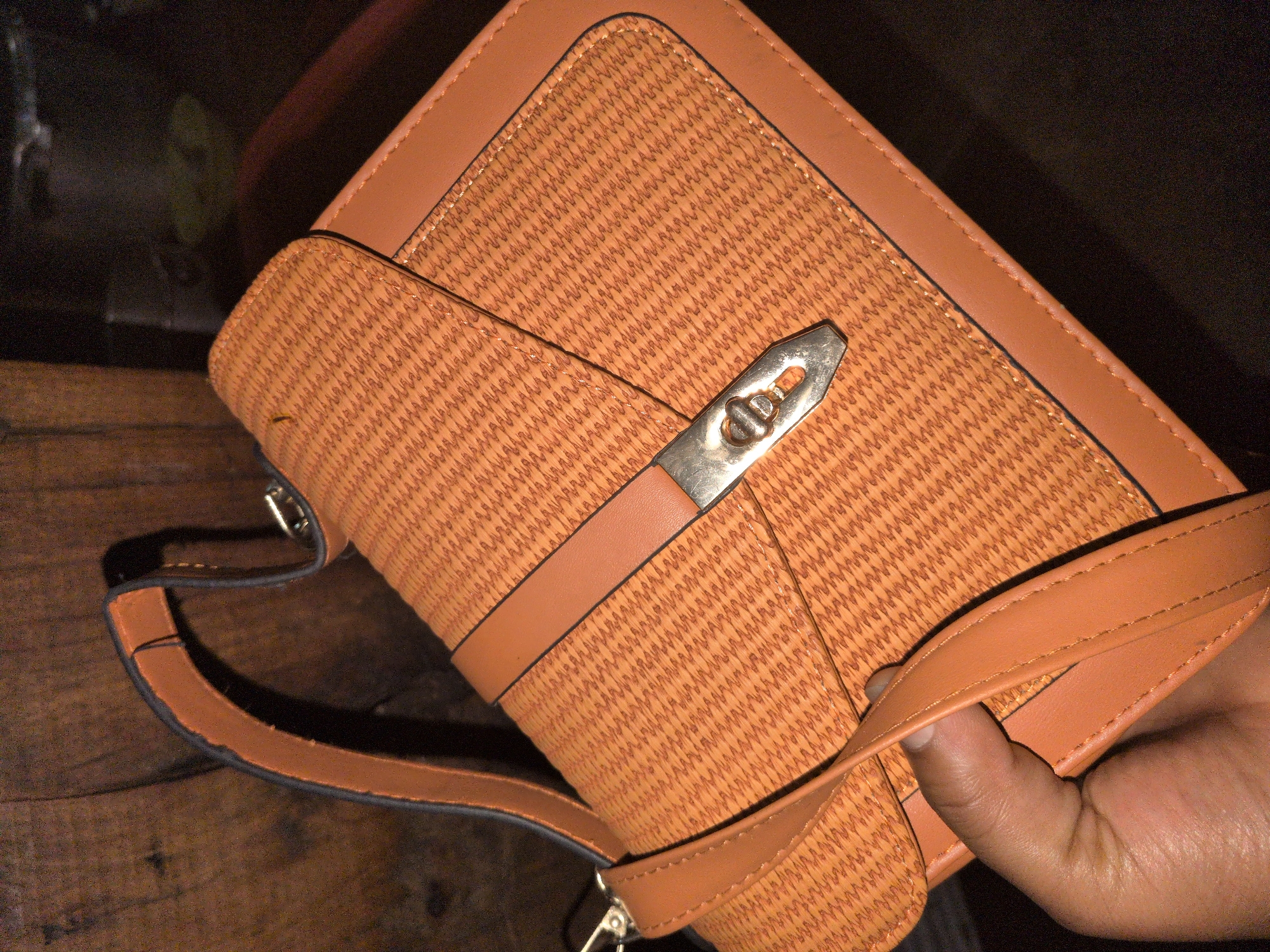 A light orange or tan women's handbag, observed in Jos, Nigeria, is positioned centrally, viewed from a slightly elevated angle. The bag features a main body and flap with a fine, repeating, textured geometric pattern, contrasted by smooth, solid-colored trim and a central horizontal band across the flap. A silver-toned metal turn-lock clasp is centrally located on the flap. A matching shoulder strap with dark, finished edges and visible stitching is attached to the bag, with a segment gripped by a hand in the lower left portion of the image. The visible hand exhibits medium-dark skin pigmentation. The background on the left is a dark brown, textured surface, consistent with wood grain. The remainder of the background is dark and out of focus, indicating low ambient light conditions, with indistinct shapes and colors faintly discernible in the distance.
