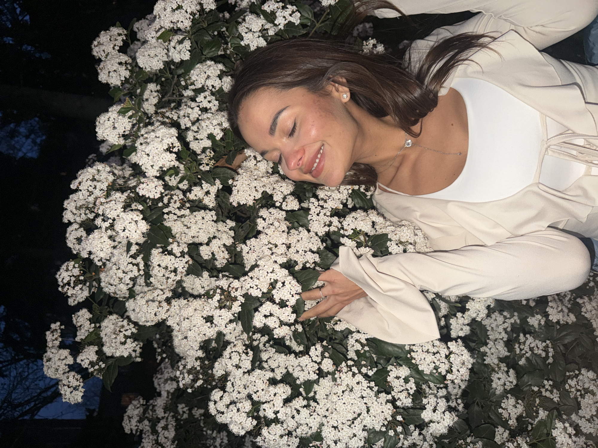 A woman is lying amongst white flowers and green leaves. She has brown hair and is wearing a white top and a beige jacket. She is smiling with her eyes closed. A necklace is visible around her neck. The scene is either at night or dimly lit. The specific location in Tbilisi, Georgia cannot be determined from the image alone.