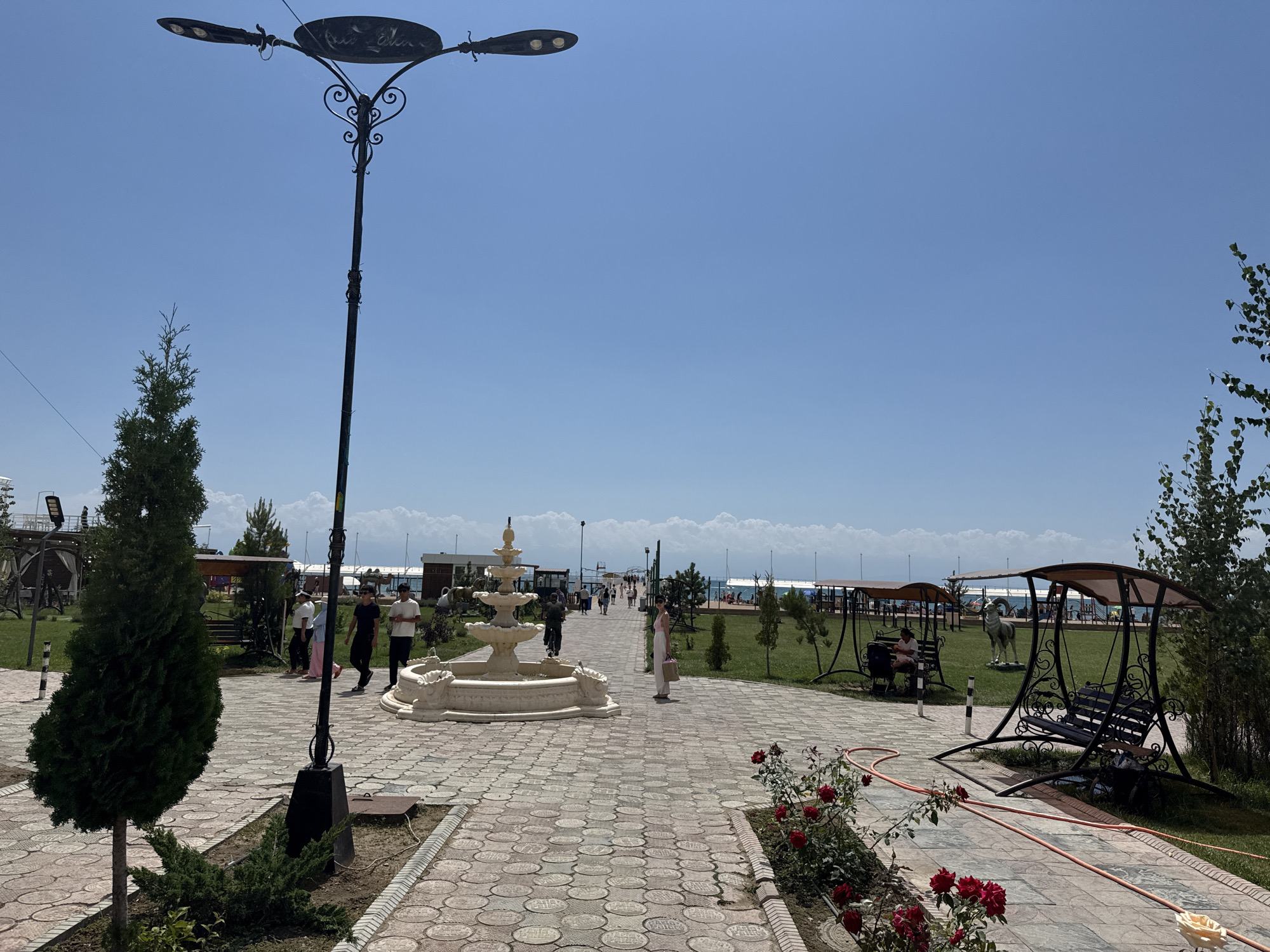 A paved walkway with patterned bricks stretches into the distance under a clear blue sky in Sary-Oy, Kyrgyzstan. On the left, a tall, dark lamppost with two lights stands beside a conical green evergreen tree. A small cluster of junipers is planted at the base of the tree. Further down the walkway, a tiered fountain with white stone tiers is visible. Several people are walking on the pathway, including a group of young men and a woman in a white outfit. To the right of the walkway, two dark metal park swings with canopies are set up on a grassy area. A statue of a ram stands between the swings. Red roses are blooming along the edge of the walkway in the foreground. In the background, more shaded seating areas and a body of water, likely Issyk-Kul Lake, are visible. The scene depicts a public park or recreational area.