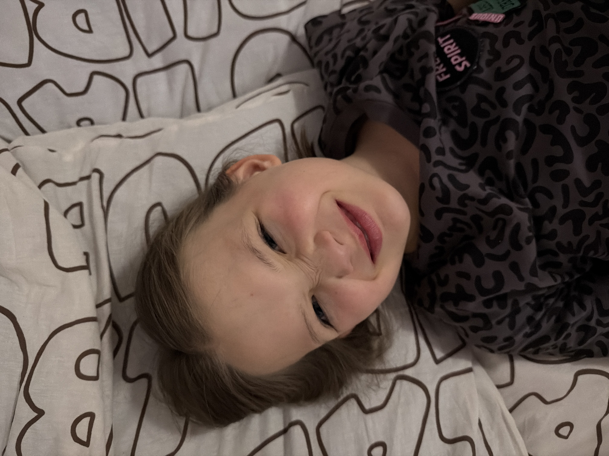 A young girl is lying on a bed with a white and brown patterned duvet cover. She is wearing a gray shirt with black leopard print and a patch that reads "Free Spirit". The location is not discernible from the image. No city or geographic information is visible.