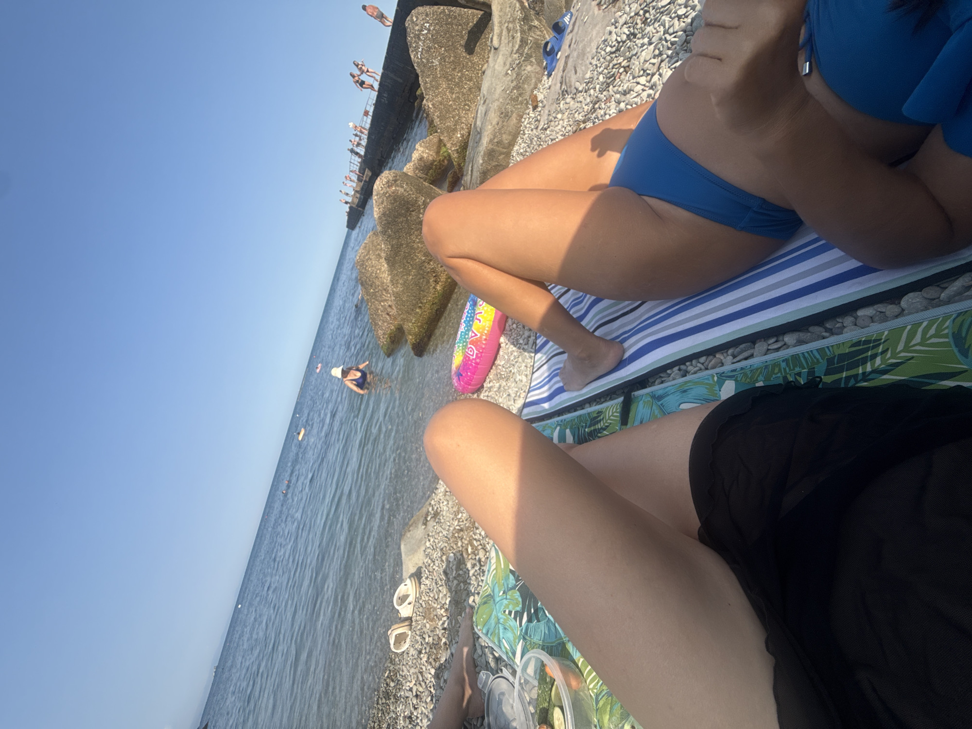 The image captures a sunny day at a beach in Partenit, Ukraine. The foreground features a close-up of two individuals lying on beach towels. One person, on the left, wears black shorts, while the other, on the right, is in a blue bikini. Scattered pebbles make up the beach surface. In the middle ground, the calm, clear sea stretches towards the horizon under a bright blue sky. Several people are visible in the water and on a pier in the distance. A colorful inflatable ring lies near the people in the foreground. The environment suggests a typical summer beach scene with people enjoying the water and sun.