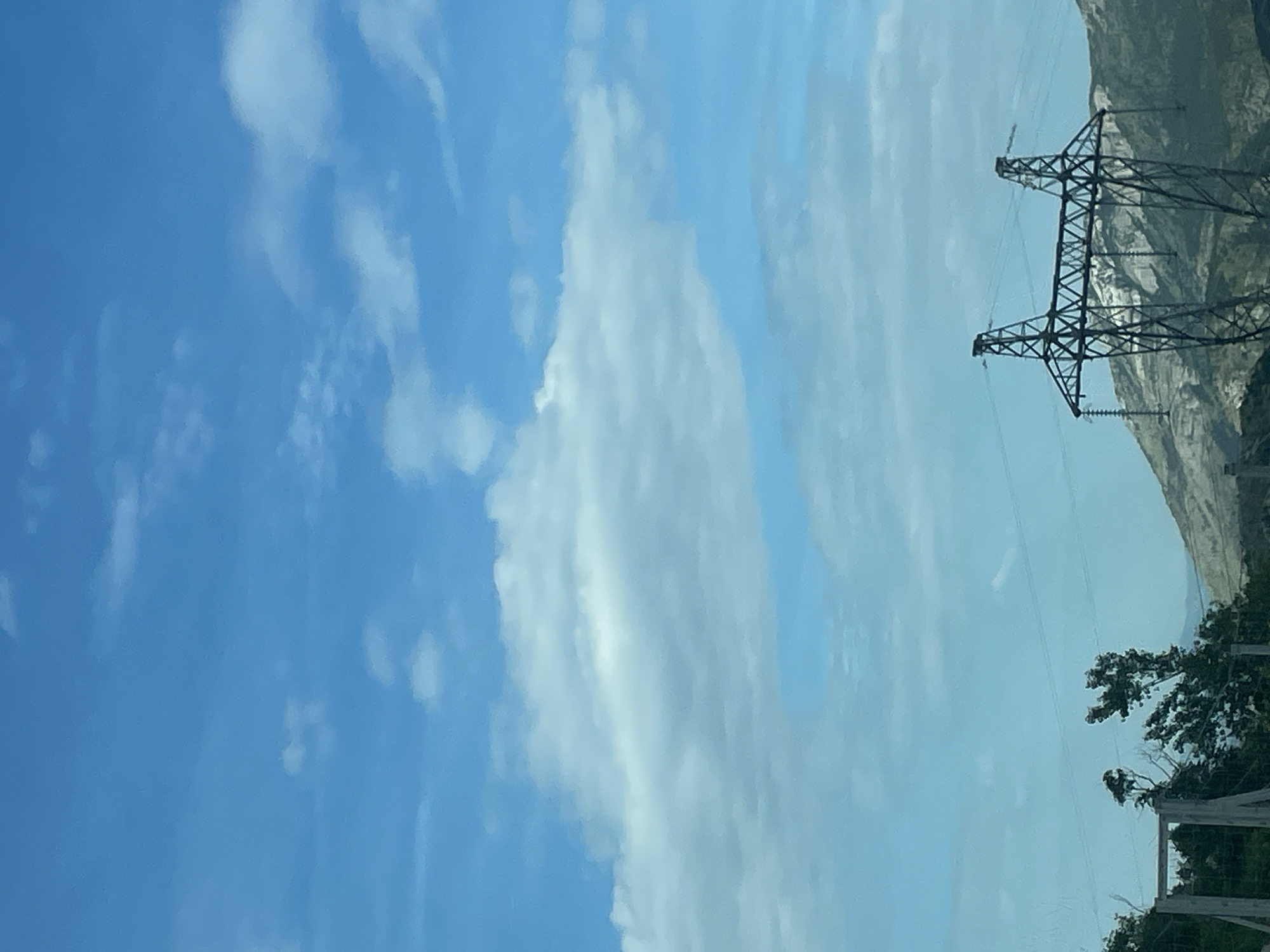 The image presents a vertically oriented view of a partially cloudy, blue sky. In the upper right, two electrical transmission towers are visible against a light-colored, rocky hillside. In the lower right corner, a tree, green foliage, and parts of a fence are present. It is impossible to say if the location is in the city of Bolesestra, Montenegro.