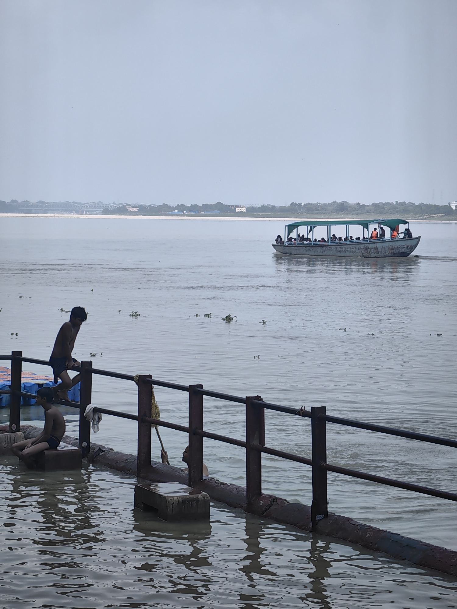 The image depicts a scene along the Ganges River in Varanasi, India. In the foreground, two young boys are visible near the water's edge. One boy, wearing dark swim trunks, is perched on a ledge, looking down. Another boy is partially submerged in the water, with only his head and shoulders visible. A metal railing separates the boys from the main body of the river.
In the middle ground, a long, narrow passenger boat is moving across the river. The boat has a green canopy and is filled with people, many wearing life vests. The river is calm, with some floating vegetation visible.
In the background, the opposite bank of the Ganges is lined with trees and buildings, suggesting an urban environment. A large bridge spans the river in the distance, and the sky is overcast. The overall atmosphere is that of a typical day on the Ganges, with both recreational and everyday activities taking place along its banks.