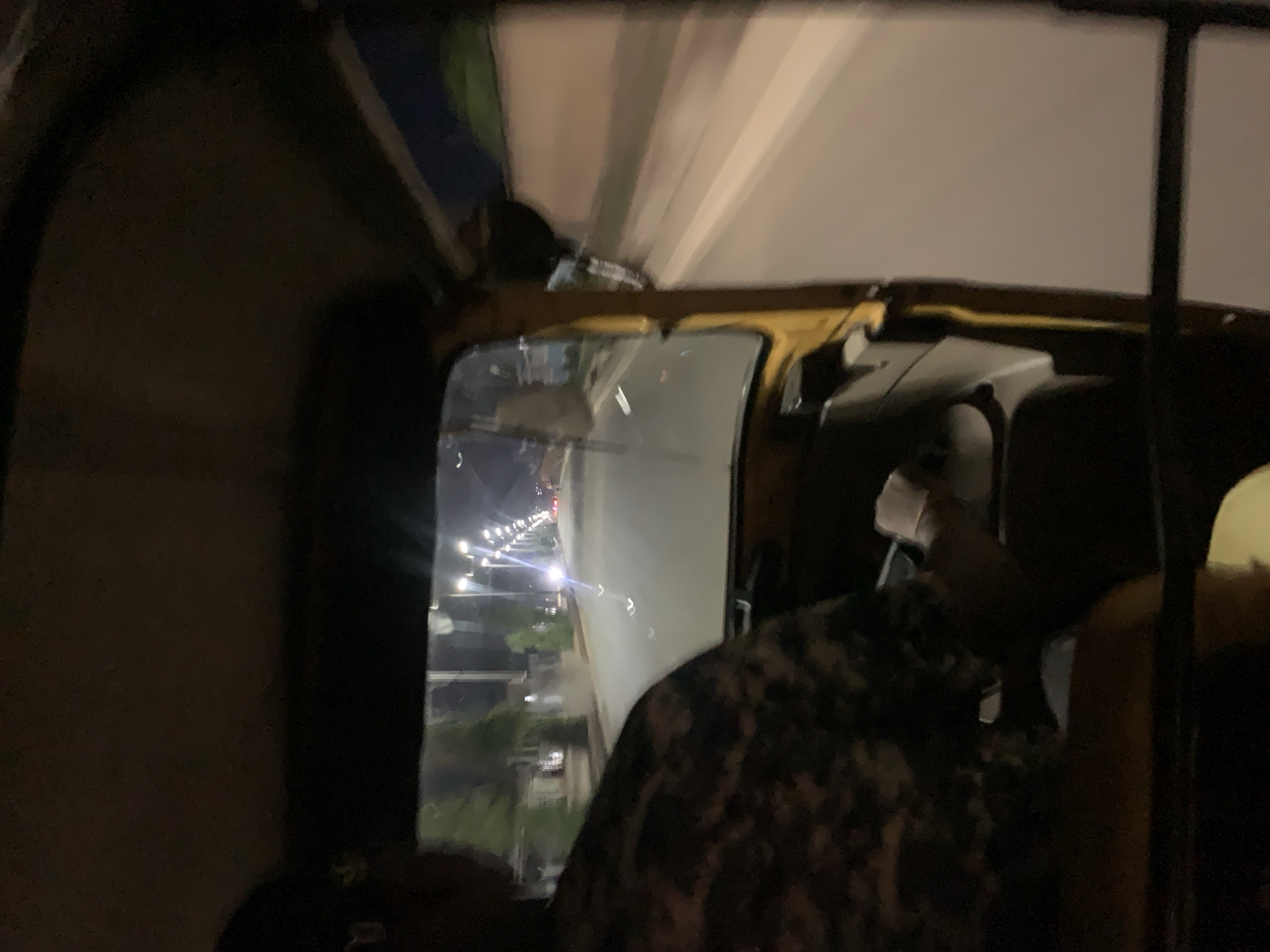 The image shows an interior view, likely from a vehicle like an auto rickshaw, traveling on a road in the city of New Stadium, Nigeria. Streetlights illuminate the road ahead, lined with trees and buildings. A person, partially visible, is wearing a camouflage-patterned garment. The scene is at night.