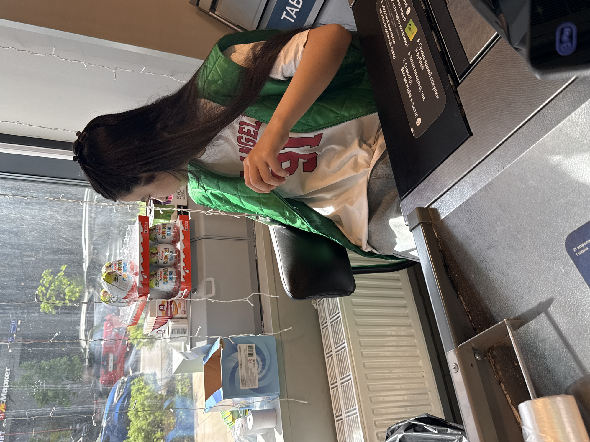 Here is a detailed description of the image:
**Location and Environment:**
* The image appears to be taken inside a small retail space or convenience store, possibly in Moscow, Russia (based on the visible Cyrillic text "Маркет").
* A window is visible on the left side of the frame, showing a view of the outdoors, including greenery, parked cars, and perhaps other buildings, suggesting an urban or suburban setting. Fairy lights are strung across the window.
* A radiator or heating unit is positioned near the till.
**People:**
* A young woman with long, dark hair is seen using a self-checkout kiosk. She is wearing a white t-shirt with a red graphic ("ANGEL 16") and a green quilted vest.
**Objects:**
* A self-checkout kiosk with a black screen displaying instructions and a payment pad is visible. The display language is in Russian.
* Kinder Surprise chocolate eggs are displayed next to the window.
* Packing tape is present near the kiosk.
**Actions and Interactions:**
* The young woman is interacting with the self-checkout kiosk, likely scanning items or making a payment.
**Overall Impression:**
The image captures a moment in a routine activity: a customer making a purchase at a self-checkout in a small retail establishment in Moscow. The scene is well-lit and provides a glimpse into everyday life.