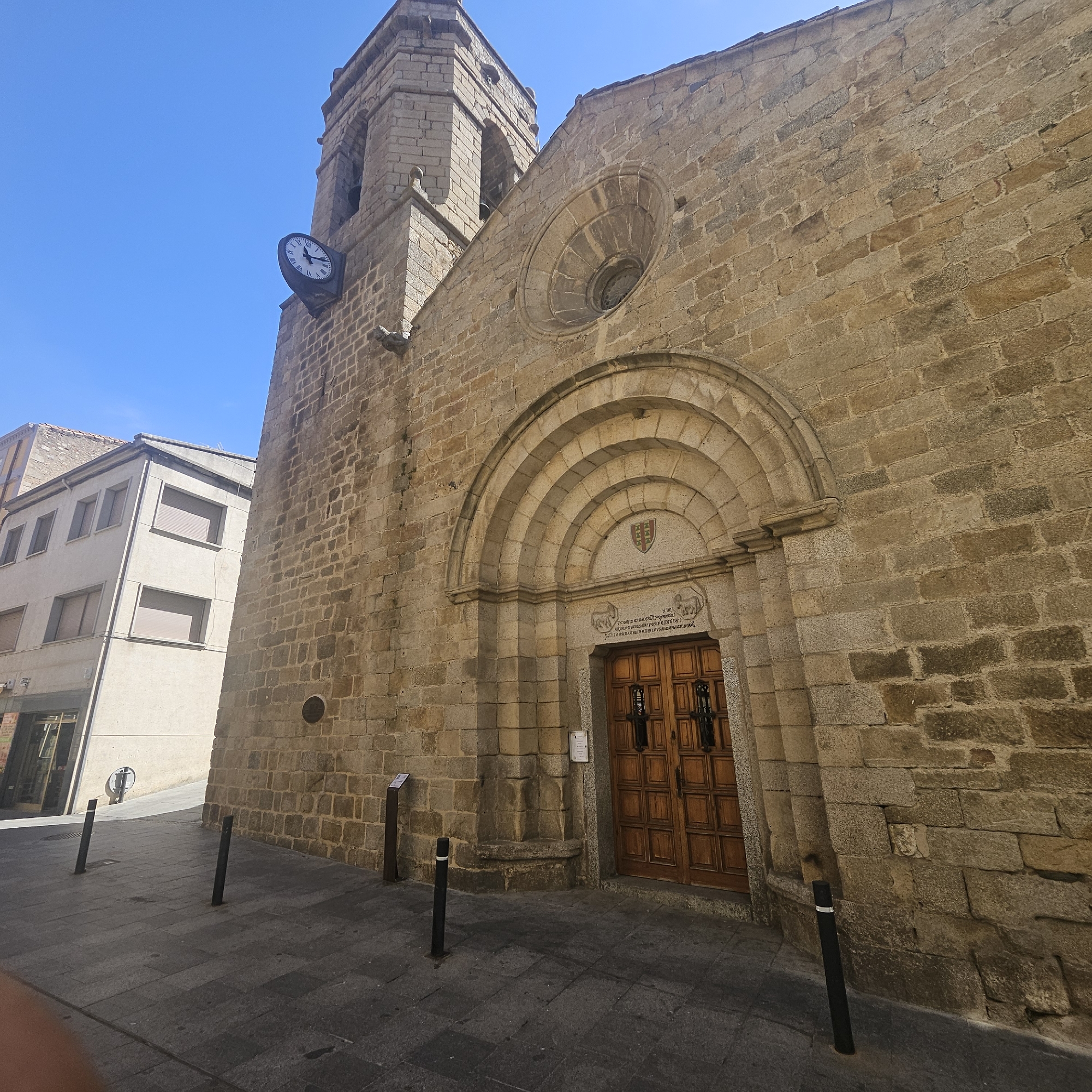 The image shows a stone church in La Jonquera, Spain, viewed from a low angle. The church has a bell tower with a clock on its side. The building is made of tan-colored stone blocks. The front facade features a large arched doorway with a wooden door and a round window above it. A three-story building with windows is visible to the left of the church tower. The foreground shows a paved street with black bollards. The sky is blue.