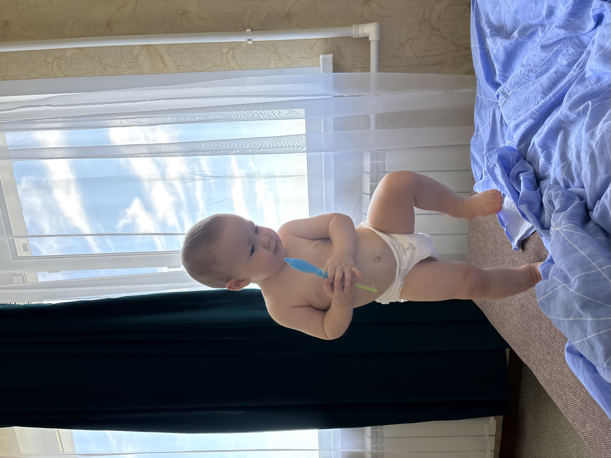 The image, taken in Naberezhnye Chelny, Russia, shows a baby wearing a diaper. The baby is lying down on a surface, possibly a bed, next to a folded blue blanket with a white geometric pattern. The baby is holding a small, blue and green object in its hands and looking towards the left of the frame with a curious expression. Behind the baby, a window is visible, with sheer white curtains allowing daylight to filter through. The curtains reveal a glimpse of a cloudy sky. A dark green curtain hangs on the left side of the window. The background wall has a textured, light-colored wallpaper. The overall lighting suggests it is daytime.