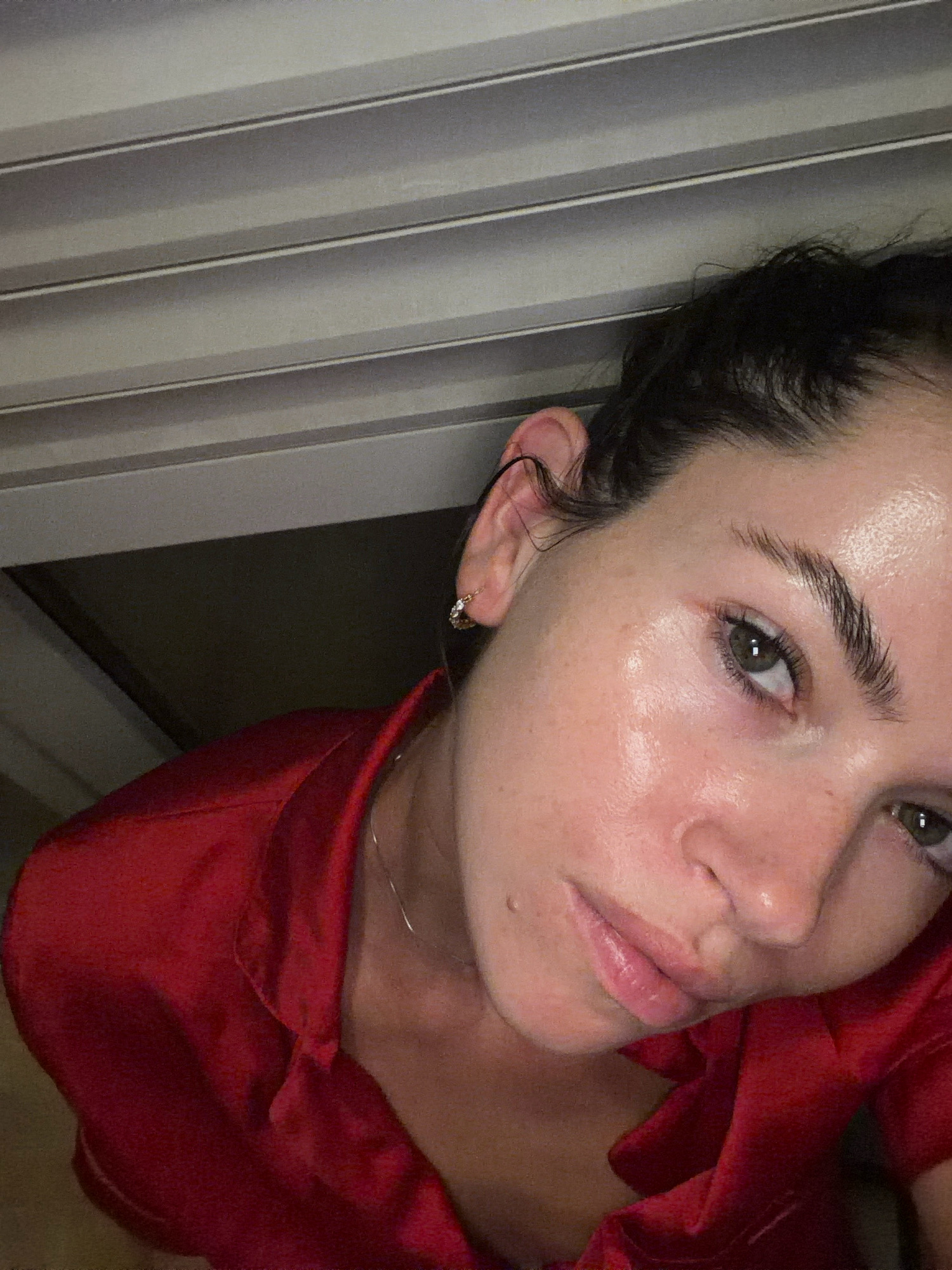 The image is a close-up selfie of a woman with dark hair and green eyes, wearing a red satin top and a small gold earring. She is looking directly at the camera. The background appears to be indoors, with horizontal blinds visible. There are no discernible indicators that this image was taken in Salou, Spain, or any other specific city. The scene is devoid of any contextual information that would allow for geographical localization.