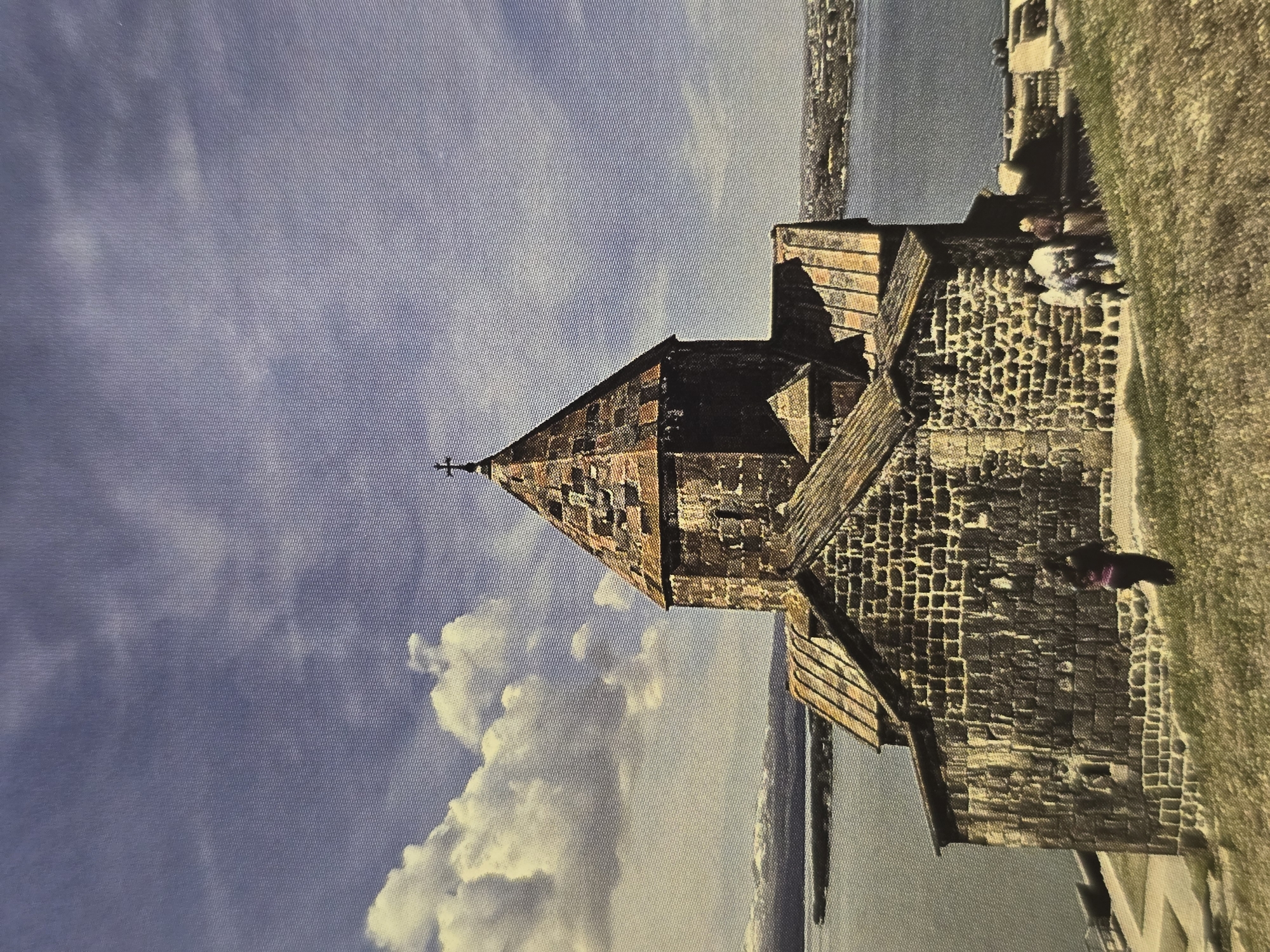 The image displays a prominent medieval stone monastery or church situated on a landmass overlooking a large body of water. The main building features robust, dark-colored stone walls and a distinctive conical dome, which is topped with a simple cross. Parts of the roof and upper sections of the structure show warmer reddish-brown stone accents, indicating a varied use of materials or weathering. The architecture suggests an ancient style, characterized by sturdy construction and unique roofing.
The setting is bright and clear, with a blue sky scattered with white, fluffy clouds, indicative of daytime. The building is positioned on a slight elevation, possibly a hill or peninsula, with a grassy area to its right. Several small figures, likely visitors or tourists, are visible on this grassy patch and near the monastery walls; one figure in a dark garment with a purple accent is more clearly seen.
The extensive body of water stretches towards the horizon, where faint outlines of what appears to be a distant city or settlement can be discerned along the shore. The overall scene conveys a sense of historical grandeur and peaceful natural beauty. There is no visible text within the image.
Location context: Sofia, Bulgaria.