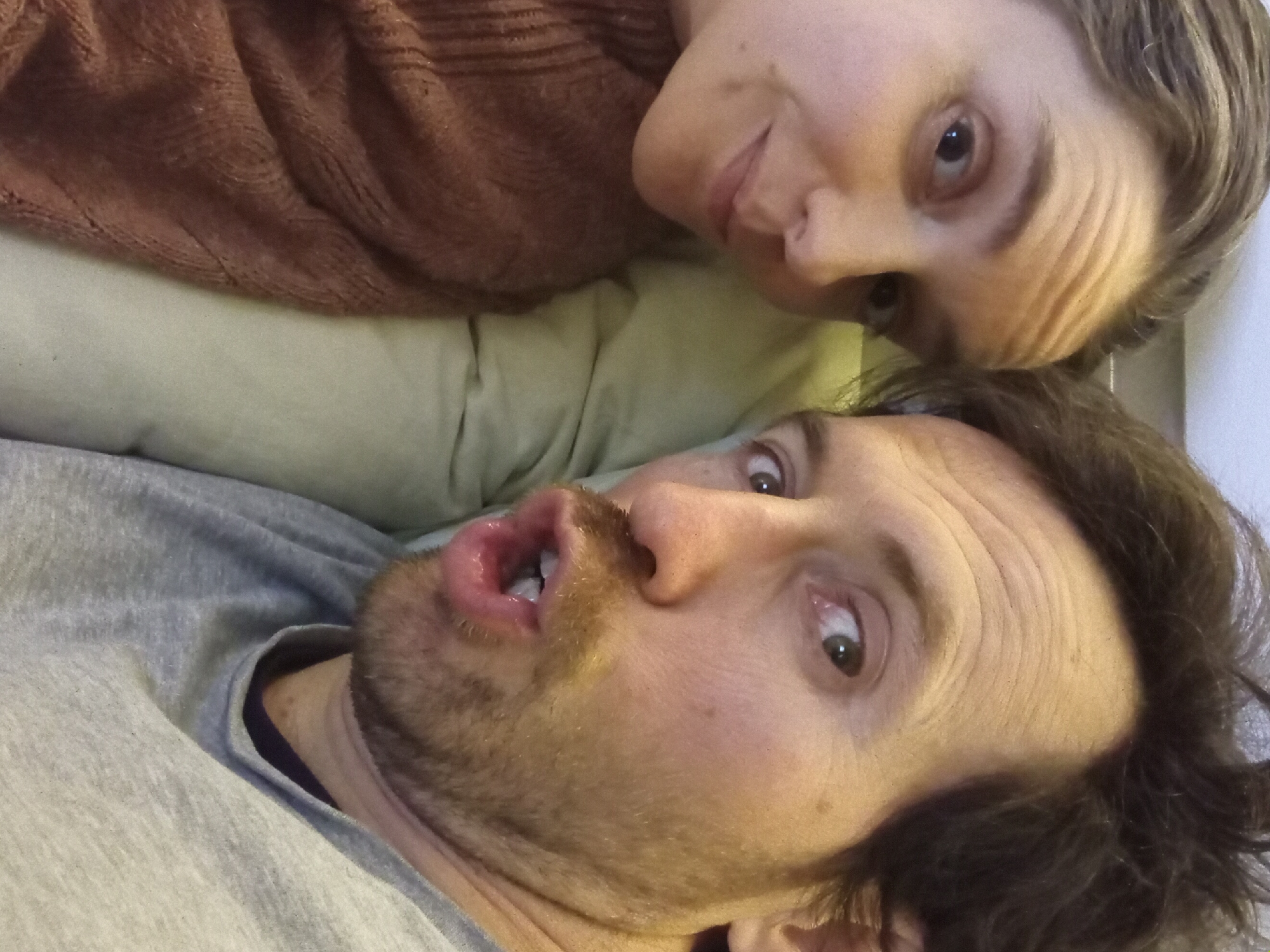 A selfie shot shows two people lying down, likely on a bed or couch, with the camera held at an angle. The person at the bottom of the frame is a man with dark hair, a light beard, and a surprised expression. He is wearing a gray shirt. Above him is a woman with light hair and a neutral expression, wearing a brown sweater. They are lying on what appears to be a light green or beige surface. The location is not discernible from the image, and there is no indication of Batumi, Georgia.