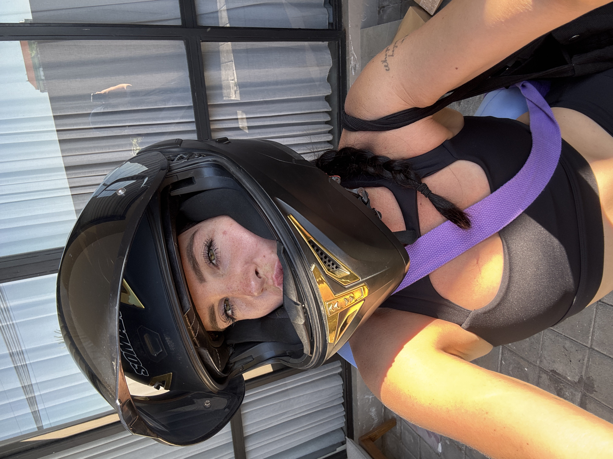 A person is wearing a black motorcycle helmet, tilted forward, obscuring most of their face except for their eyes, nose, and mouth. The helmet has gold accents. The person is wearing a black sports bra with a purple strap. Part of their arm and shoulder are visible, and there appears to be a tattoo on their arm. The background shows a window with blinds and a glimpse of what might be a wall or fence. The lighting suggests it is daytime, possibly outdoors, with sunlight casting shadows. The image is oriented vertically.