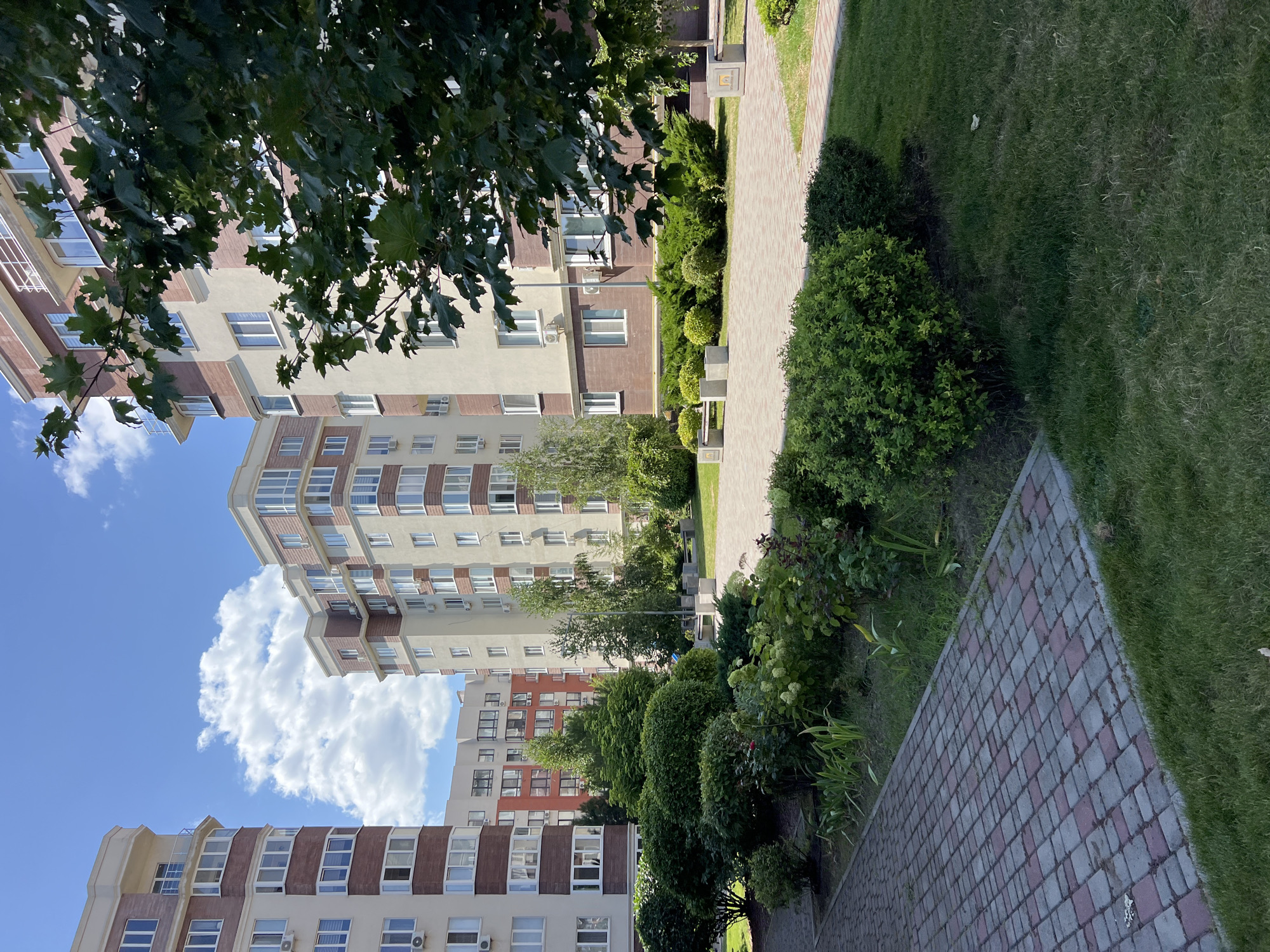 The image captures a sunny day in Kryzhanivka, Ukraine, showcasing a modern residential complex with its well-maintained courtyard. Multiple apartment buildings, characterized by their beige and brick-red facades with white window frames and balconies, dominate the background. Lush greenery, including manicured bushes, trees, and a flower bed, forms a vibrant foreground and midground. A paved walkway, laid with reddish-pink and grey interlocking stones, leads the viewer's eye through the scene. Benches and concrete cube-shaped seating are visible in the courtyard area, suggesting a communal space for residents. The sky is a clear blue, adorned with fluffy white clouds, and a large tree branch with green leaves partially obscures the view of one of the buildings on the right. The overall impression is one of a peaceful and aesthetically pleasing urban living environment.