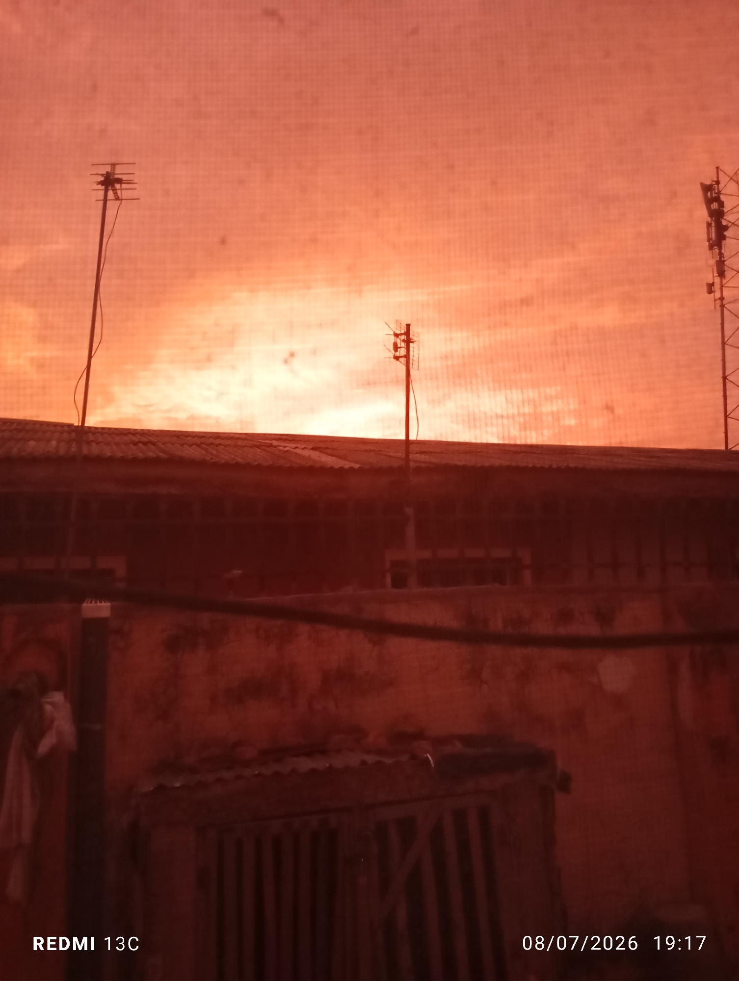 The image captures a sunset or sunrise over a residential area, likely in Obada, Nigeria, given the context of the query. The sky is filled with a warm, orange hue, suggesting atmospheric conditions common during twilight. In the foreground, the roof of a building is visible, appearing to be made of corrugated metal. Several antennas are mounted on poles, one on the left and another on the right side of the frame, which could be for television or communication purposes. Below the roofline, the details of the building's facade are dark and indistinct, but suggest a weathered texture. A small, enclosed structure with a slatted door is present in the lower portion of the image, and a piece of white cloth hangs on the left side. The bottom of the image displays text: "REDMI 13C" and "08/07/2026 19:17", indicating the device used for capture and the date and time. The overall scene is dimly lit, with the vibrant sky being the brightest element.