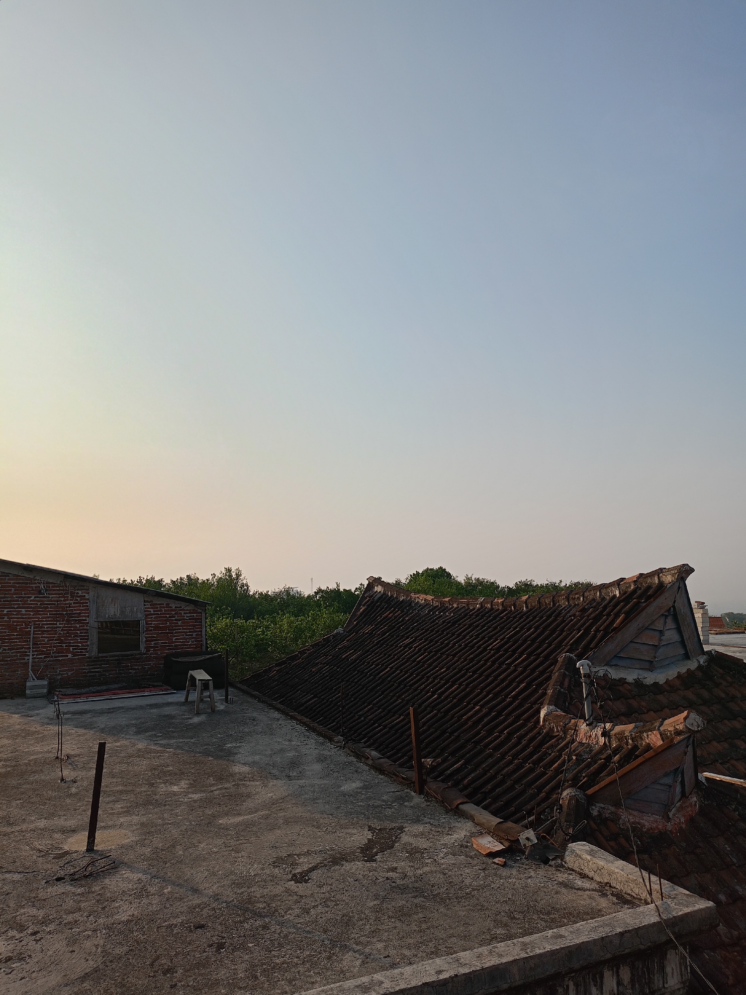 The image captures a rooftop view in Batu, Indonesia, during what appears to be late afternoon or early evening. The sky is a pale, clear blue, fading to a warmer yellow-orange hue near the horizon. In the foreground, a concrete rooftop is visible, showing signs of wear and tear with some debris and metal poles protruding from the surface. To the left, a weathered brick building with a corrugated metal roof extension is partially visible. Further to the right, an older tiled roof with an intricate, curved design dominates the mid-ground, showing signs of age and disrepair, with some tiles missing and exposed wooden structural elements. Beyond the rooftops, a dense line of green trees forms a natural barrier, suggesting the presence of a less urbanized area or a park. The overall impression is one of a quiet, somewhat aged urban or semi-urban environment on the outskirts of Batu, bathed in the soft light of the setting sun. No people are present in the scene.