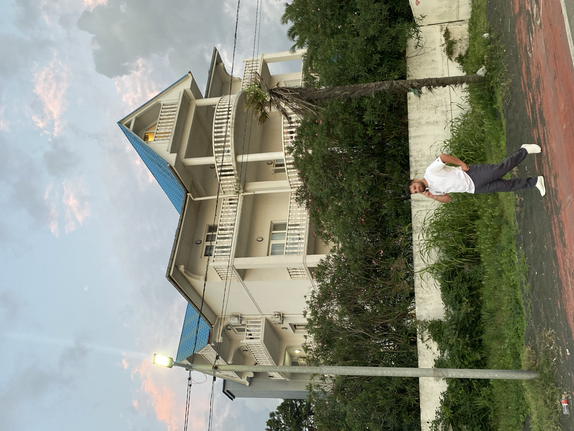 In Ureki, Georgia, a man in a white t-shirt and grey pants walks along a path, talking on his phone. He wears white sneakers. Behind him, a white building with a blue roof stands amidst lush green foliage. A street lamp is visible to the left of the building. The sky is overcast with hints of pink and orange clouds.