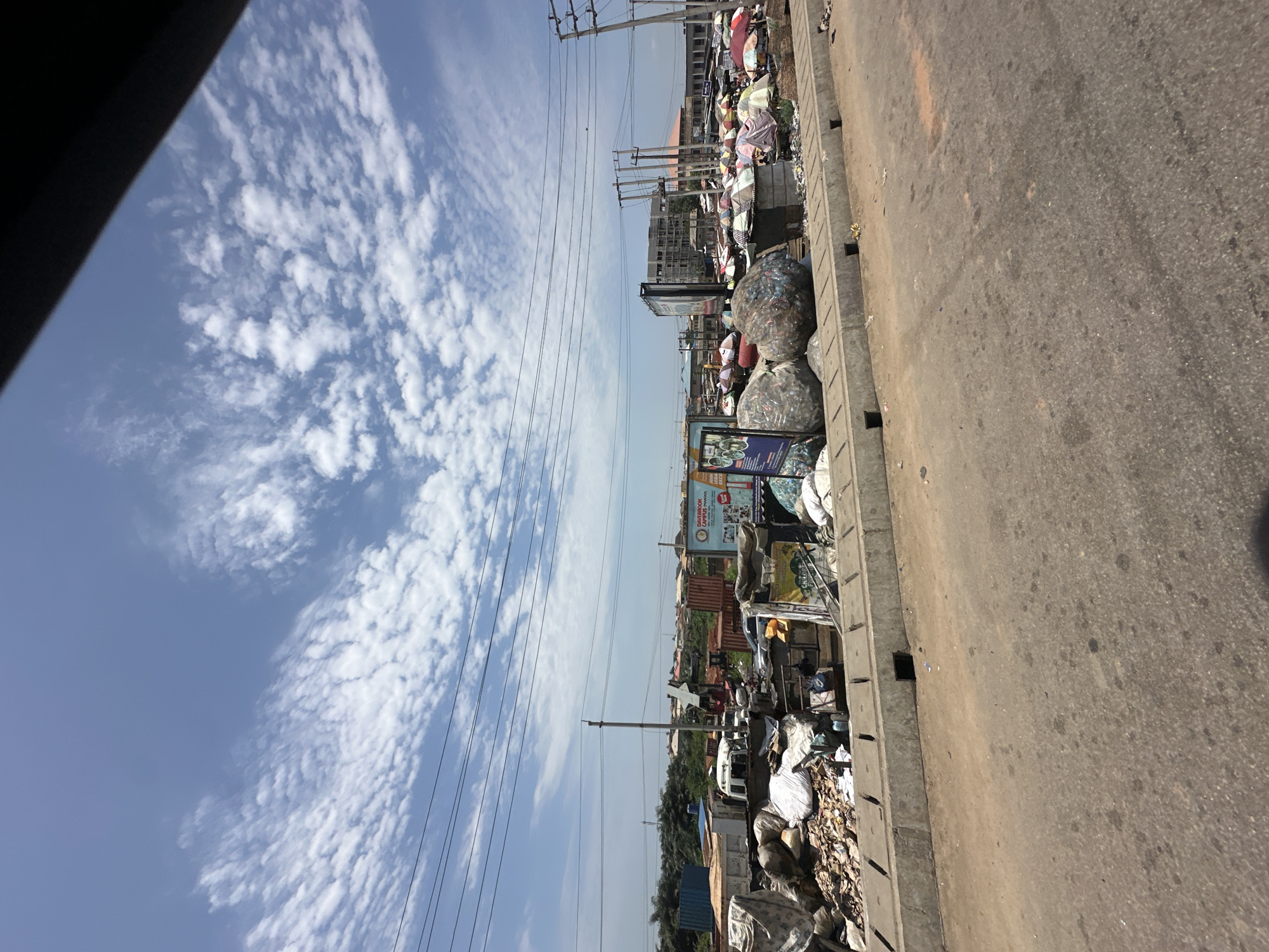This image depicts an outdoor scene in Magboro, Nigeria, characterized by a considerable amount of waste and debris piled along a roadside. The foreground shows a section of a road and a concrete barrier with several square openings. Behind the barrier, a large accumulation of various types of refuse is visible, including plastic bottles, bags, fabric, and other unidentifiable waste materials. Some large, compressed bales of plastic are prominent in the midground, along with scattered smaller piles of garbage.
In the background, there are several structures that suggest an urban or semi-urban environment. These include shipping containers, trucks, a crane, and buildings, some of which appear to be under construction. Numerous colorful umbrellas are set up, indicating a market or commercial area, likely situated amongst or adjacent to the waste. Electrical poles and power lines are visible, as are trees and other vegetation further in the distance. The sky above is blue with scattered, white, fluffy clouds. There are no visible people in the image. The overall impression is one of an active, albeit cluttered, area with evidence of both commerce and significant waste management challenges.