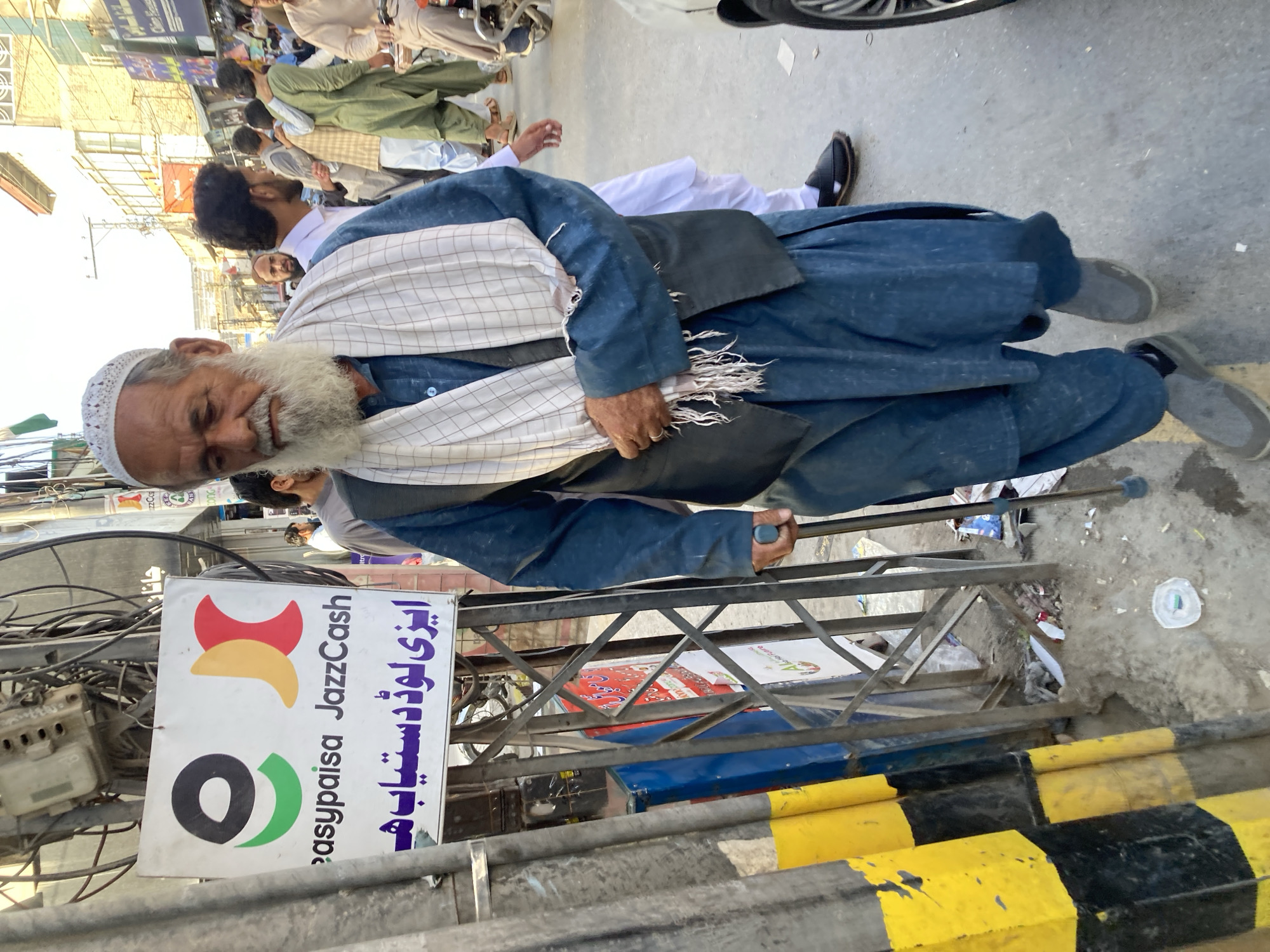 In Quetta City Tehsil, Pakistan, an elderly man with a white beard and a white skullcap walks along a street using a cane. He is dressed in a blue shalwar kameez and wears a checkered scarf. A sign for "easypaisa JazzCash" is visible next to him. The street is crowded with people and there are vehicles parked on the side. The road is marked with yellow and black painted barriers. Overhead wires crisscross the scene.
