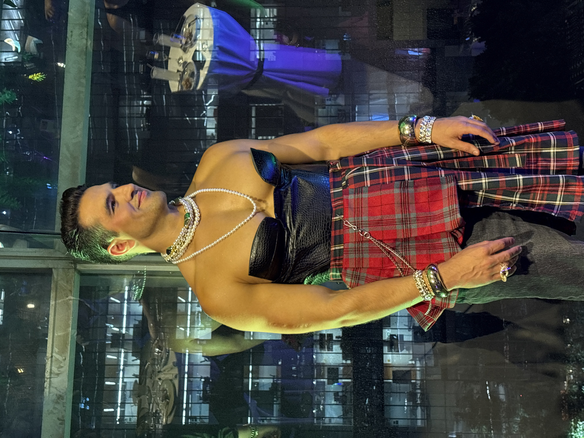 The image captures a person wearing a black corset-style top, a plaid skirt, pearl necklaces, bracelets, rings, and dark jeans. The setting is a dimly lit interior with a reflective surface creating a mirrored effect. Through a window, the illuminated cityscape of Mexico City, Mexico is visible.