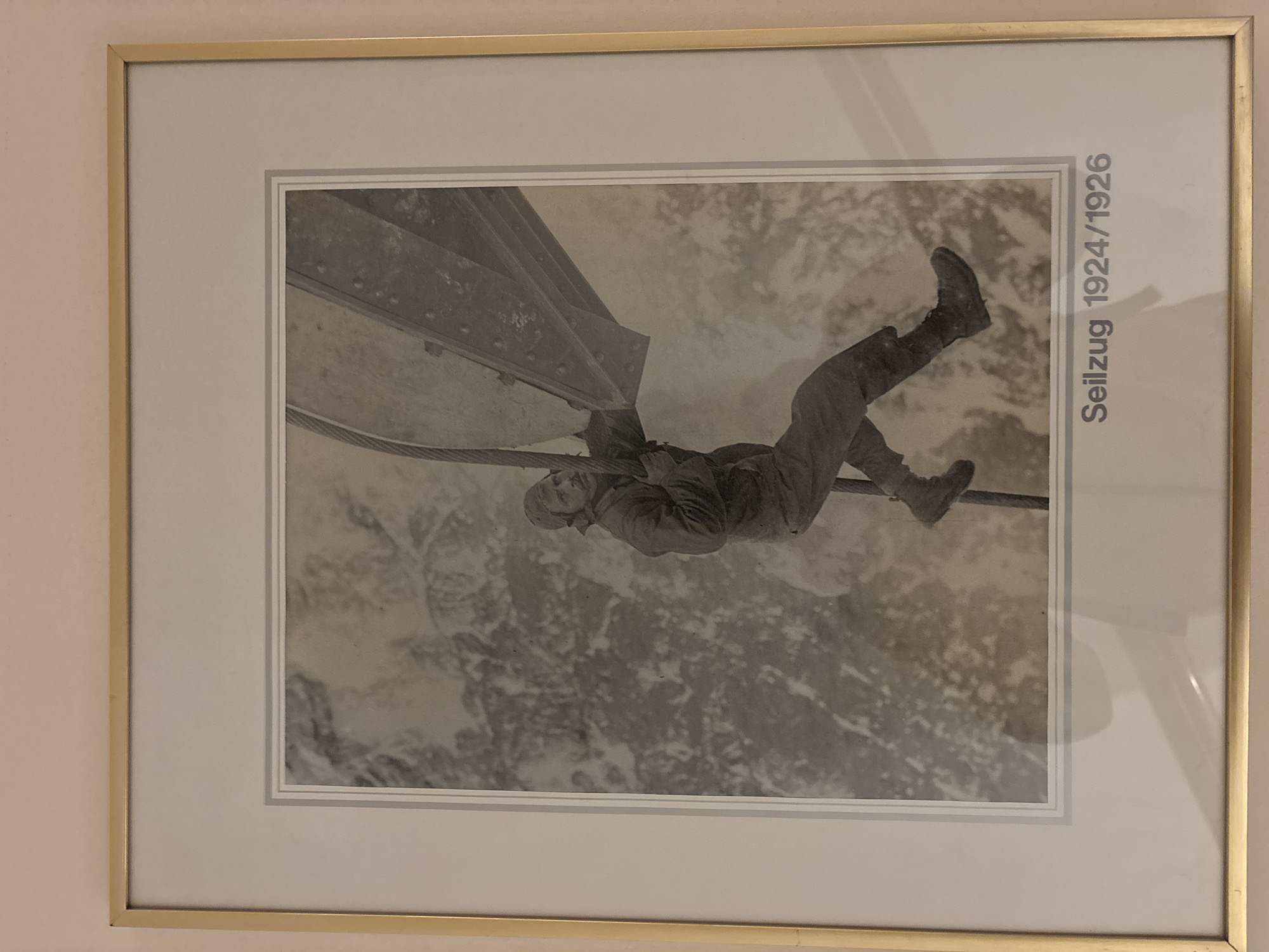 The image is a framed black and white photograph showing a man climbing on a thick cable. He is wearing outdoor clothing including a hat and heavy boots. He is holding onto the cable with his hands and has his legs wrapped around it. In the background, there is a blurry, mountainous landscape covered in snow. The photograph is titled "Seilzug 1924/1926" which translates to "Cable Car 1924/1926". This title, combined with the snowy mountainous terrain, suggests the photograph was taken in a high-altitude location, possibly in connection with the construction or operation of a cable car system. Given that the title refers to a period between 1924 and 1926 and mentions "Seilzug" which can also refer to a cable railway, it is plausible that this depicts an early stage of a cable transport system in Ehrwald, Austria, as Ehrwald is known for its mountainous terrain and cable car facilities.