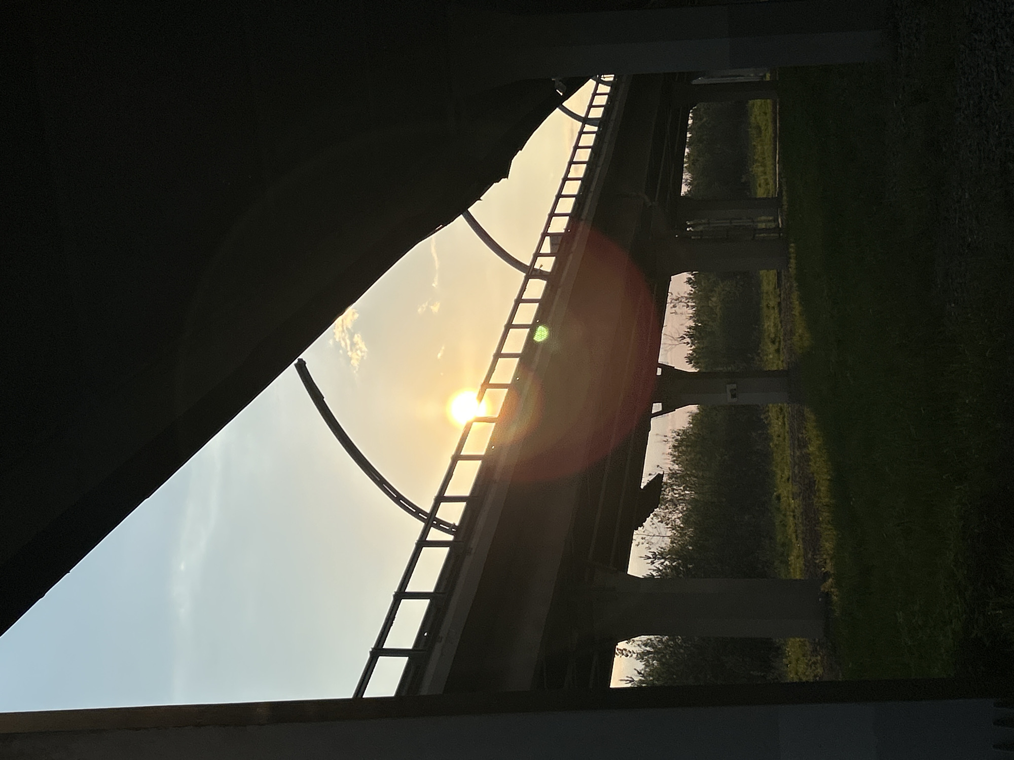 The image shows a view from under a bridge, looking upwards and outwards. The sun is directly above and obscured by the bridge supports, creating a lens flare effect. The sky is visible through the gaps in the bridge structure. Below the bridge, vegetation is visible. The location is within the city of Saint Petersburg, Russia, based on the presence of bridges in the area. The image contains no visible people, actions, or interactions. The environment consists of concrete structures, sky, and vegetation.