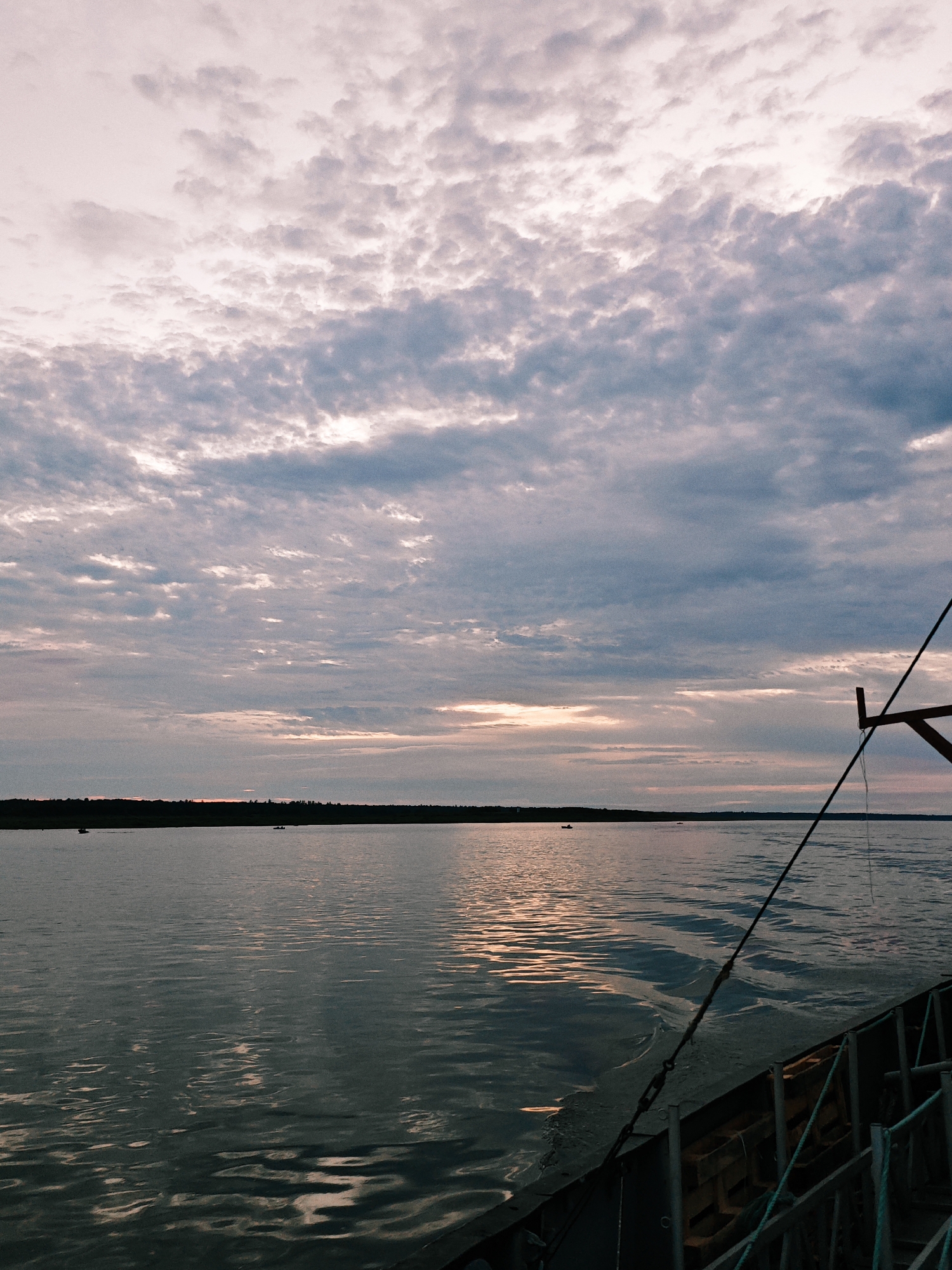 This media file depicts a serene lake scene at dusk in Nikulino, Russia. The sky is a canvas of dramatic clouds, painted in hues of pale pink, grey, and muted blue, suggesting either dawn or sunset. The sun is low on the horizon, casting a warm, golden glow that reflects on the calm surface of the lake.
The water is gently rippling, creating a mesmerizing play of light and shadow. In the distance, a dark, tree-lined shore forms a stark contrast against the vibrant sky and water. A few small boats are scattered across the lake, their occupants likely enjoying the peaceful evening.
In the foreground, a portion of a boat or ferry is visible, hinting that the photo was taken from another vessel. A thick rope, secured by metal links and hooks, stretches diagonally across the frame, adding a sense of depth and perspective. The presence of wooden crates and metal railings suggests it's a working craft, perhaps a ferry transporting people or goods across the lake.
The overall mood is tranquil and contemplative, evoking a sense of peace and the beauty of nature. The time of day is clearly indicated by the soft light and the colors of the sky, characteristic of twilight. There are no visible text elements in the image.