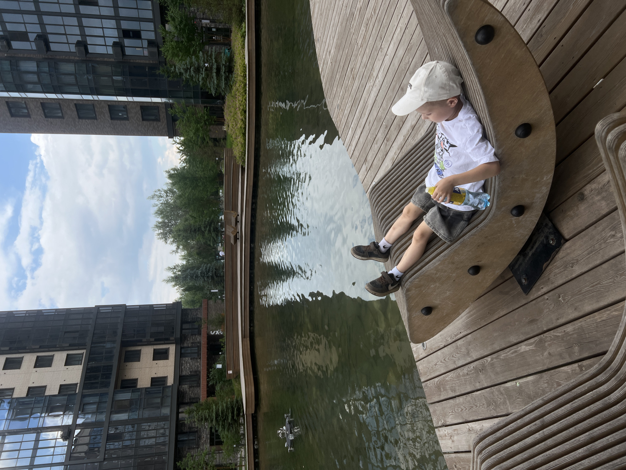 In an outdoor urban setting, likely within the city of Krasnoyarsk, Russia, a young boy is relaxing on a wooden, rocking lounge chair. He is lying on his back, wearing a white t-shirt with a colorful graphic, dark shorts, and a white baseball cap. He holds a bottle of water in his right hand. The boy is positioned on a wooden deck that surrounds a body of water. In the water, a small drone is visible. Behind the water, there are modern, multi-story buildings with dark glass facades, suggesting a contemporary urban development. Lush green trees and landscaping are present between the buildings and the water. The sky is partly cloudy.
