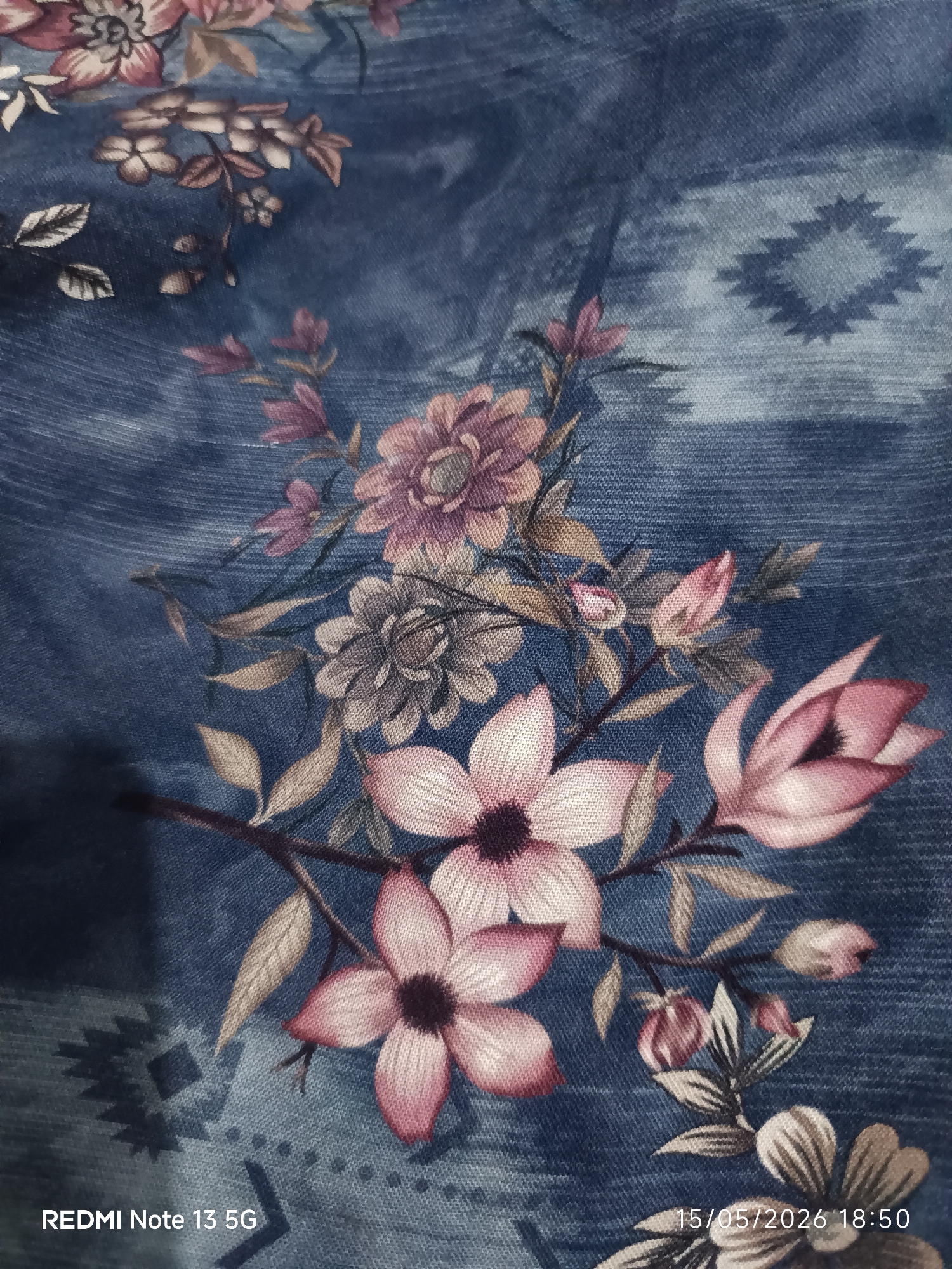 The image is a close-up shot of a fabric with a floral and geometric pattern. The fabric appears to be a denim-like material, dyed in shades of blue. Overlaid on this blue background is a pattern consisting of various flower designs in colors ranging from pink to mauve, along with neutral-toned leaves and stems. Interspersed between the floral motifs are geometric shapes resembling diamonds or stylized stars, also in a lighter shade of blue. The photograph was taken with a Redmi Note 13 5G camera, as indicated by the watermark in the bottom left corner. The date and time stamp, "15/05/2026 18:50," are present in the bottom right corner. There is no indication of the location Tanjung Morawa, Indonesia visible in this image.