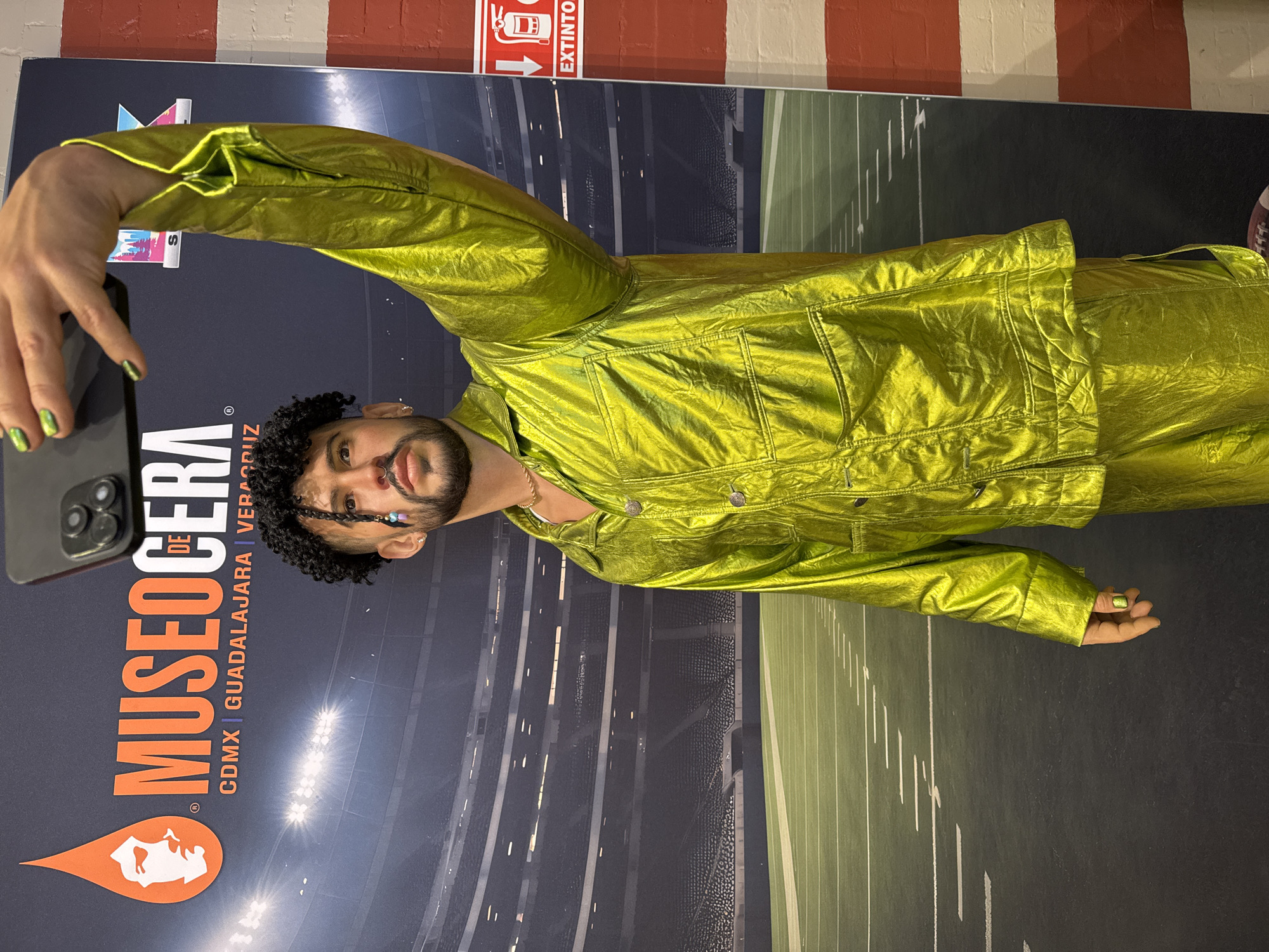 A person wearing a shimmering lime-green jacket and pants, likely a wax figure of Bad Bunny, is posing for a selfie in front of a backdrop for the Museo de Cera, a wax museum with locations in Mexico City, Guadalajara, and Veracruz, Mexico. The museum's logo, an orange droplet containing a white head, is visible. The backdrop depicts a sports stadium with stadium lights. A red and white painted wall with a fire extinguisher sign is visible in the background.