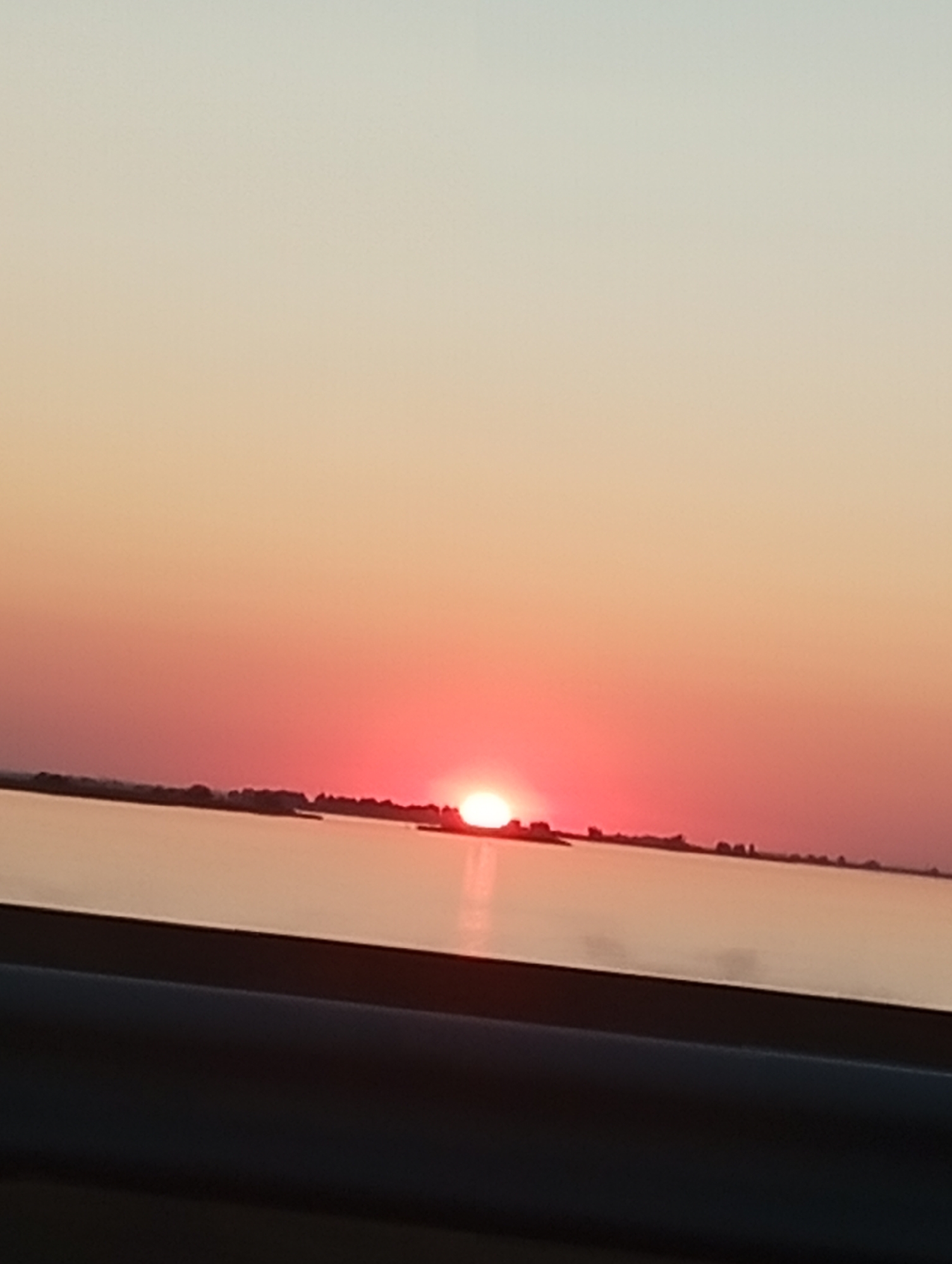 A sunset over a body of water is captured from a slightly elevated perspective, possibly from a vehicle. The sun, a bright orb of light, is partially obscured by a thin, dark horizon line that appears to be land, perhaps with some silhouetted structures or trees. The sky transitions from a pale, almost white hue at the top to a vibrant pink and red closer to the horizon, reflecting on the calm water. In the foreground, a dark, blurry element, likely the interior of a car or a railing, occupies the bottom portion of the frame, suggesting the photo was taken through a window or from a moving platform. The overall impression is one of a serene, natural landscape at dusk.